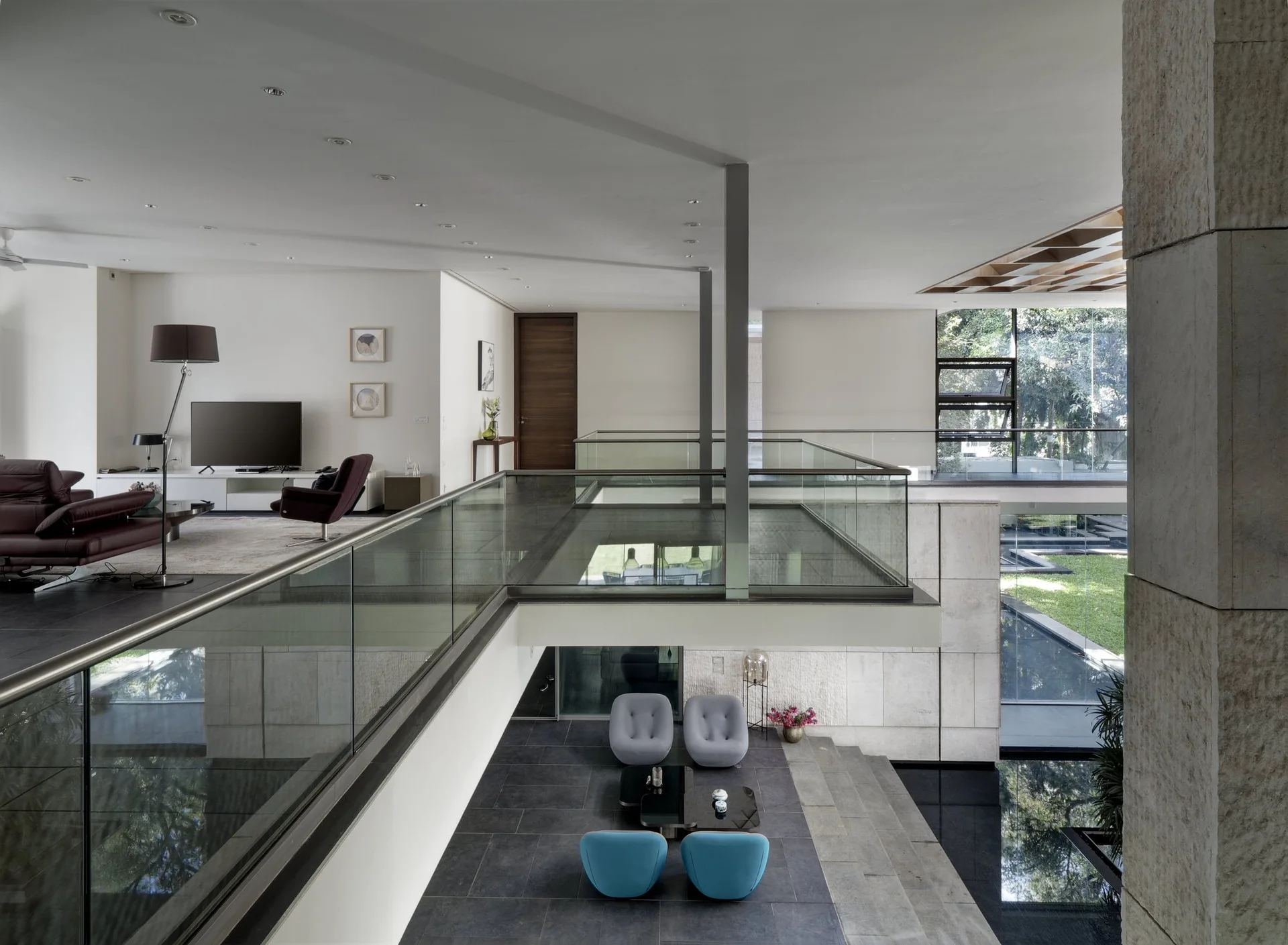 A modern split-level interior with a glass-railed mezzanine overlooking a lower lounge area with blue armchairs and an indoor water feature.