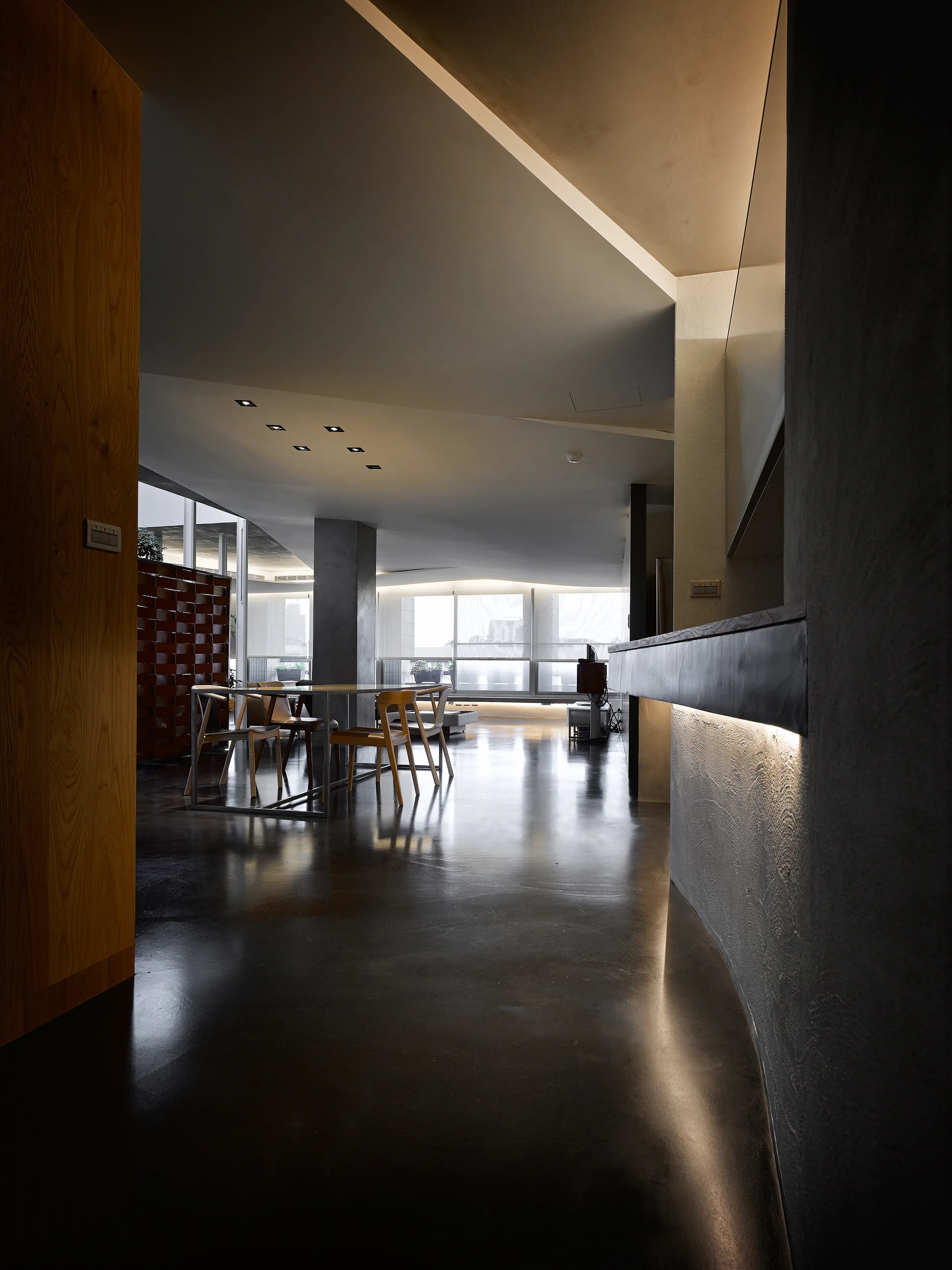 A minimalist dining room features a wooden table and chairs on dark polished floors under angular ceilings with warm integrated cove lighting.