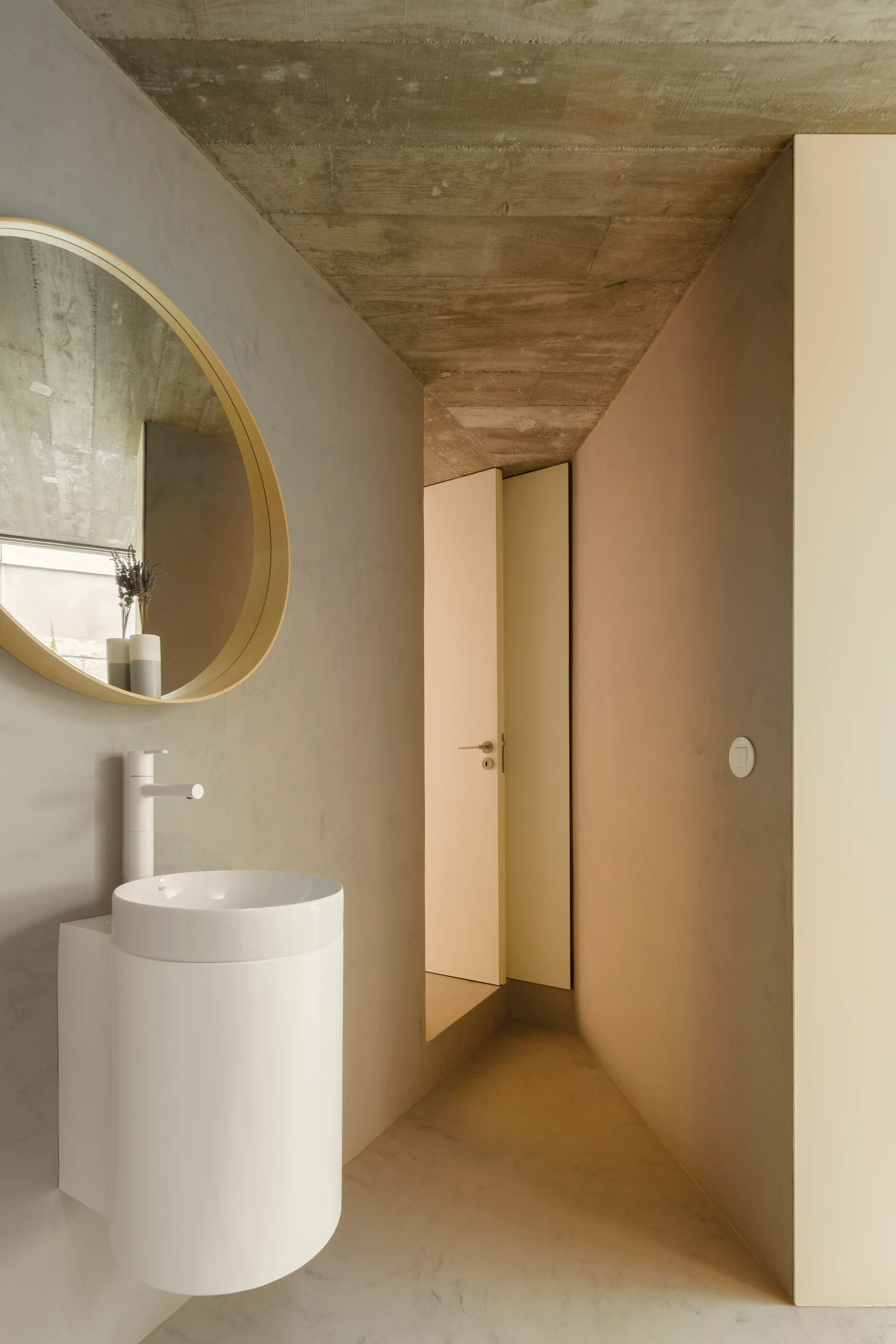A minimalist powder room features a white cylindrical sink, a circular wood-framed mirror on a plaster wall, and an exposed raw concrete ceiling.