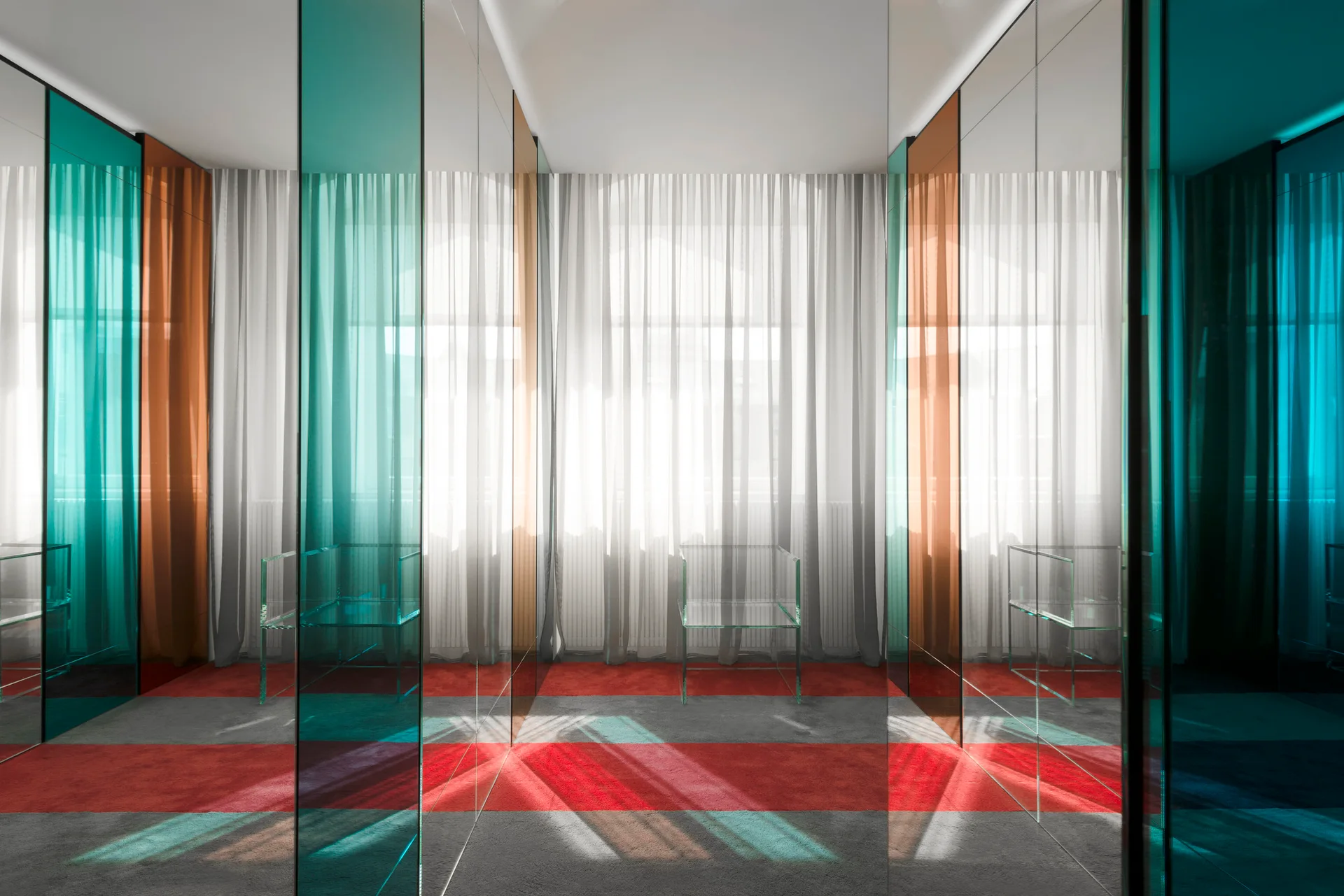 A minimalist room features teal and copper colored glass partitions, sheer white window curtains, and a striped red and gray carpet with transparent chairs.