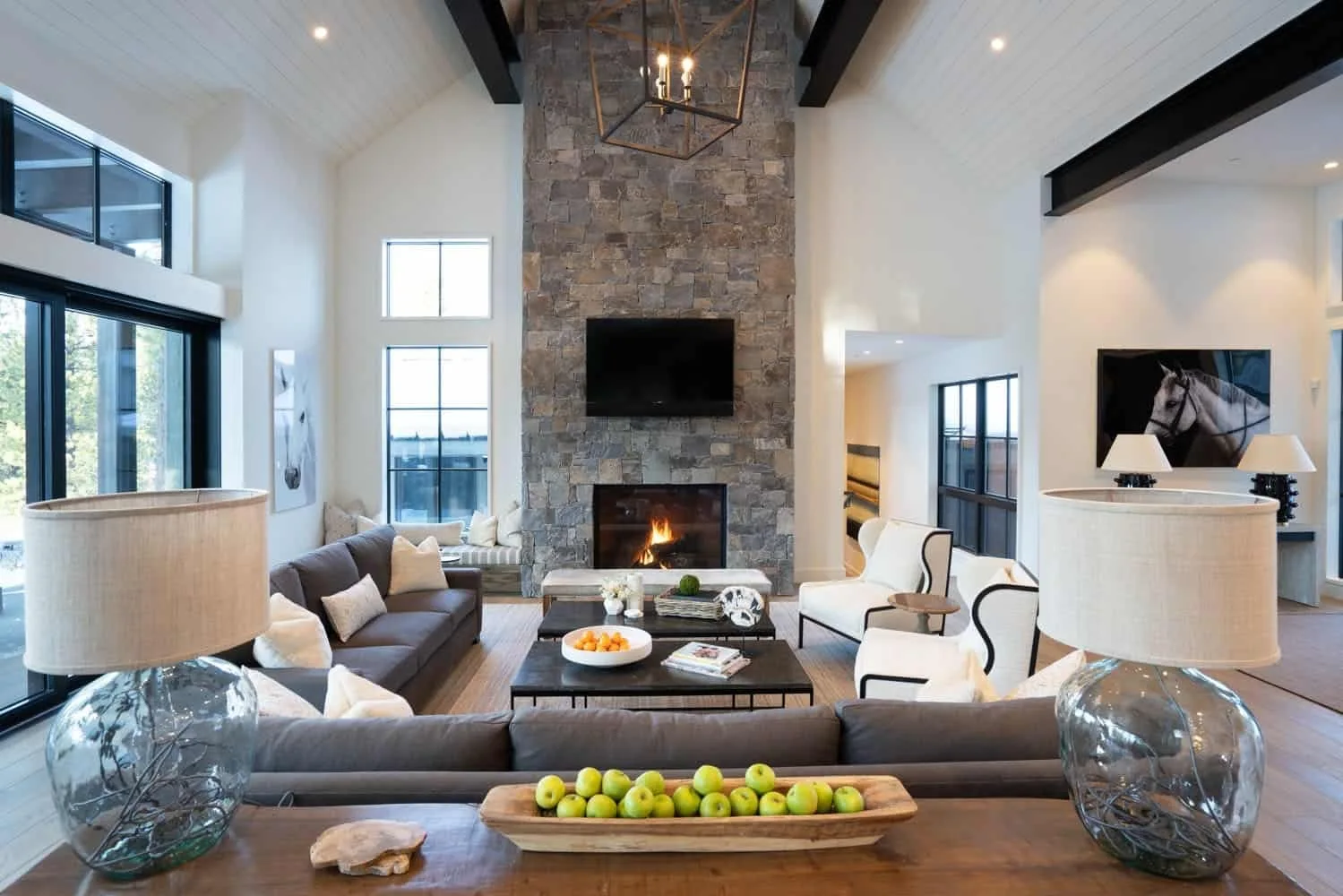 A spacious living room features a tall stone fireplace, a vaulted ceiling with dark wood beams, and grey and white seating arranged around low coffee tables.