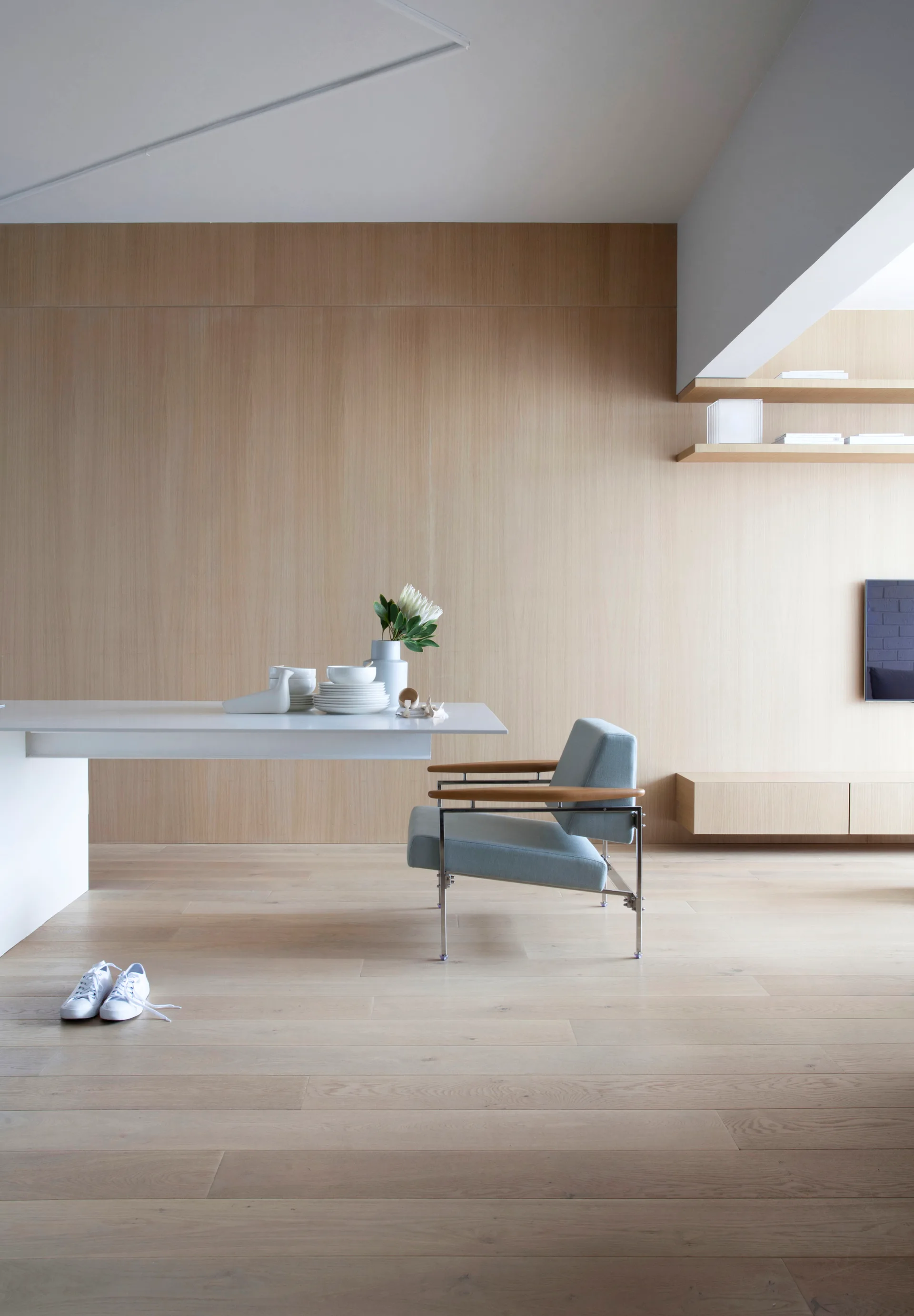 A minimalist room featuring light wood wall paneling, a white cantilevered table with a pale blue armchair, and floating wooden shelves.