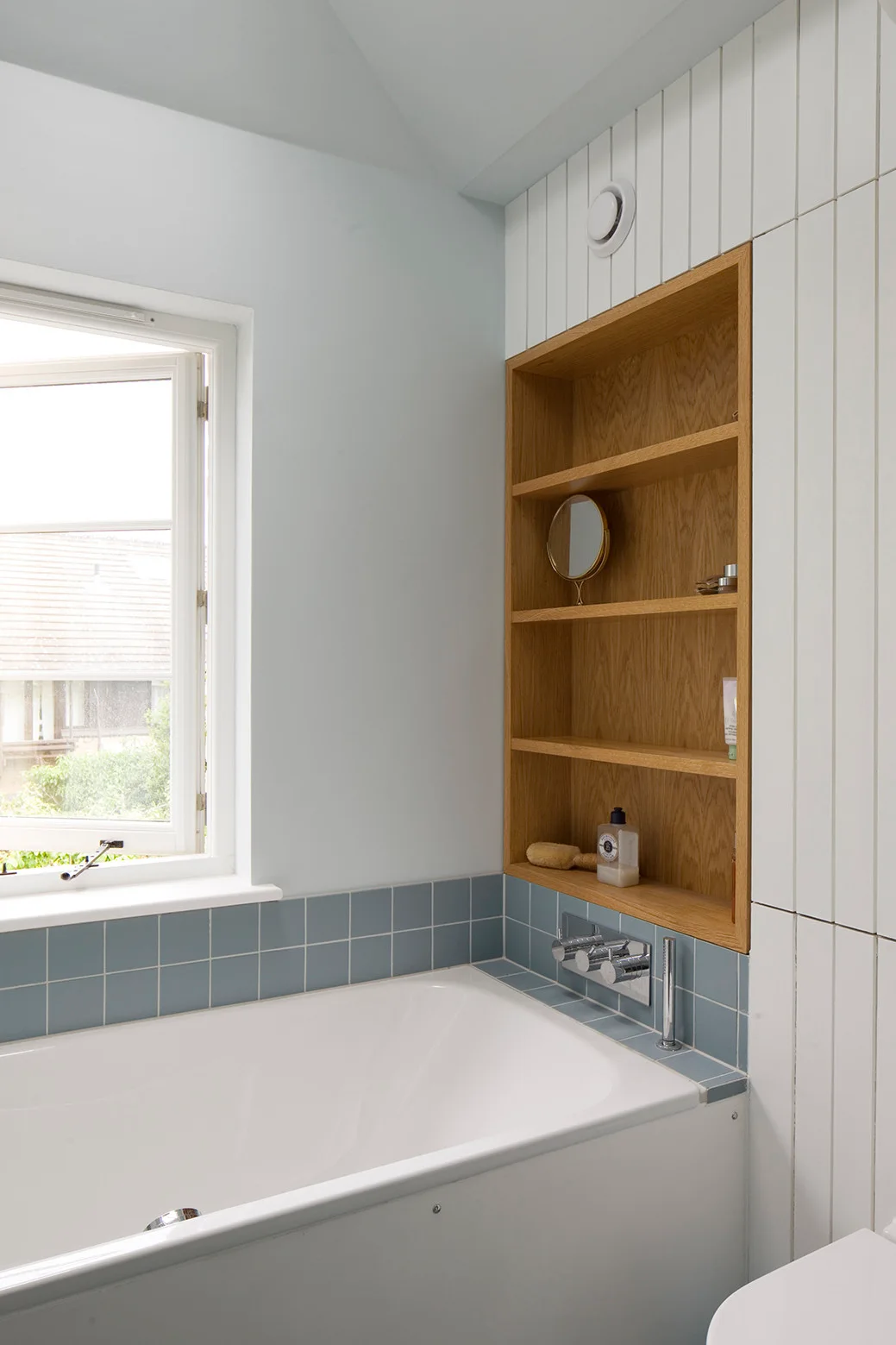 A bathroom featuring a white bathtub, light blue tiled trim, vertical white wall paneling, and a built-in light wood shelf niche.