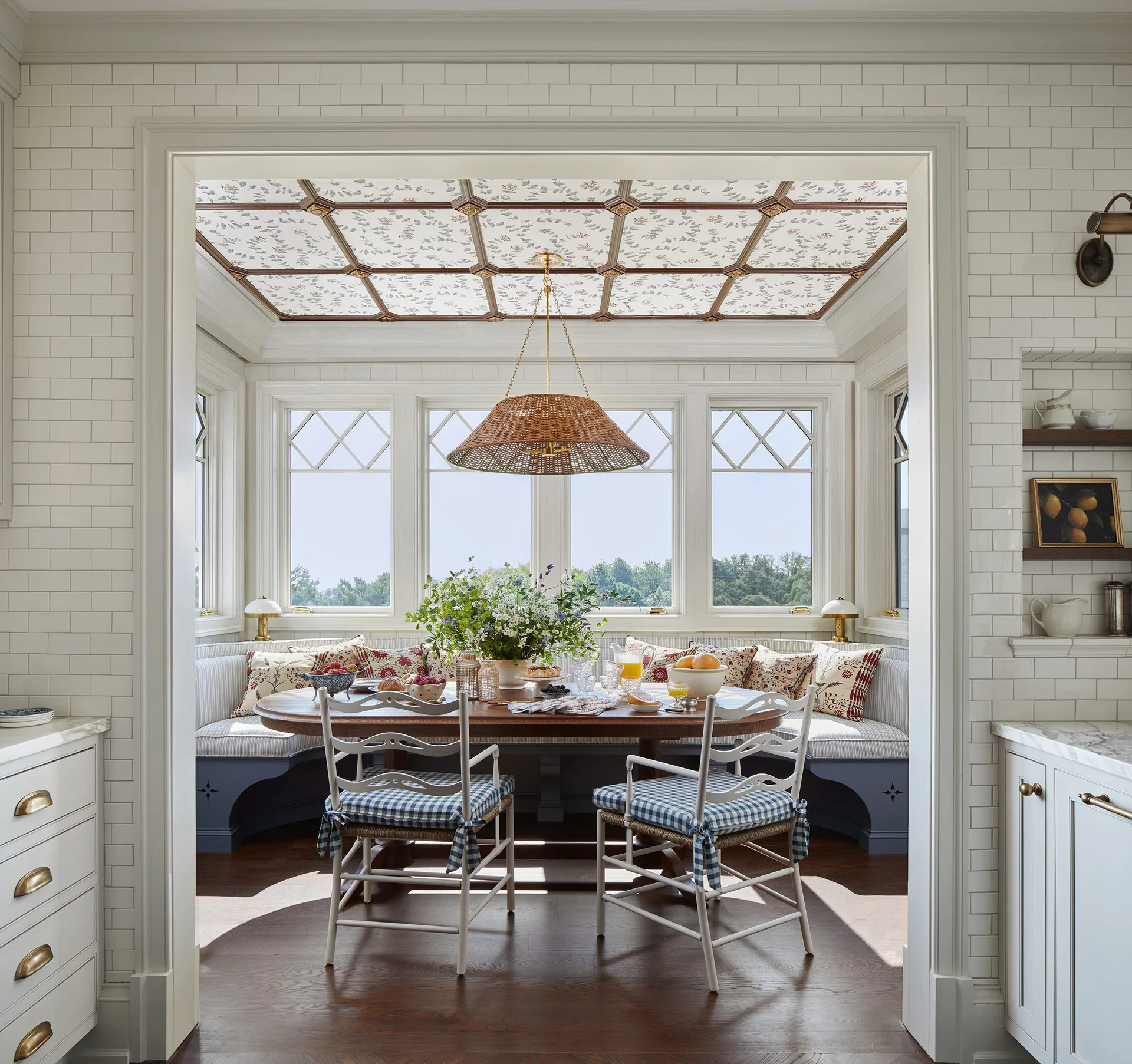A breakfast nook featuring a blue built-in banquette, wood table, and a coffered ceiling with floral wallpaper, viewed through a tiled entryway.