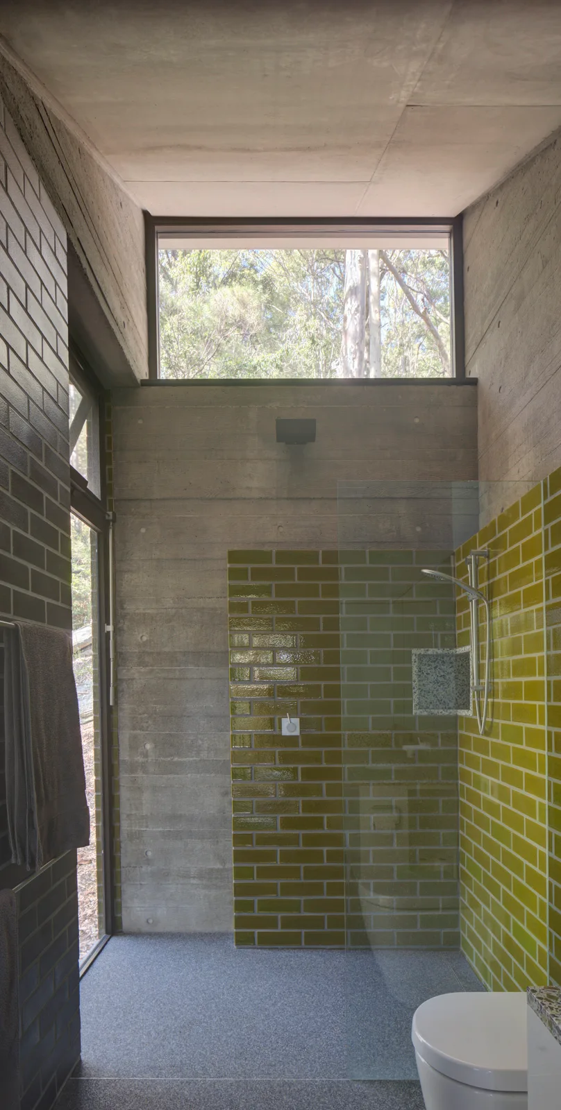 A minimalist bathroom with a glass walk-in shower, glossy olive green subway tiles, raw concrete walls, and a high window looking out to trees.