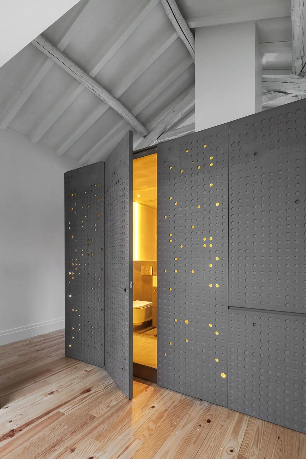 A dark grey perforated partition door partially opens to reveal a warm yellow-lit bathroom with a toilet under a sloped beam ceiling.