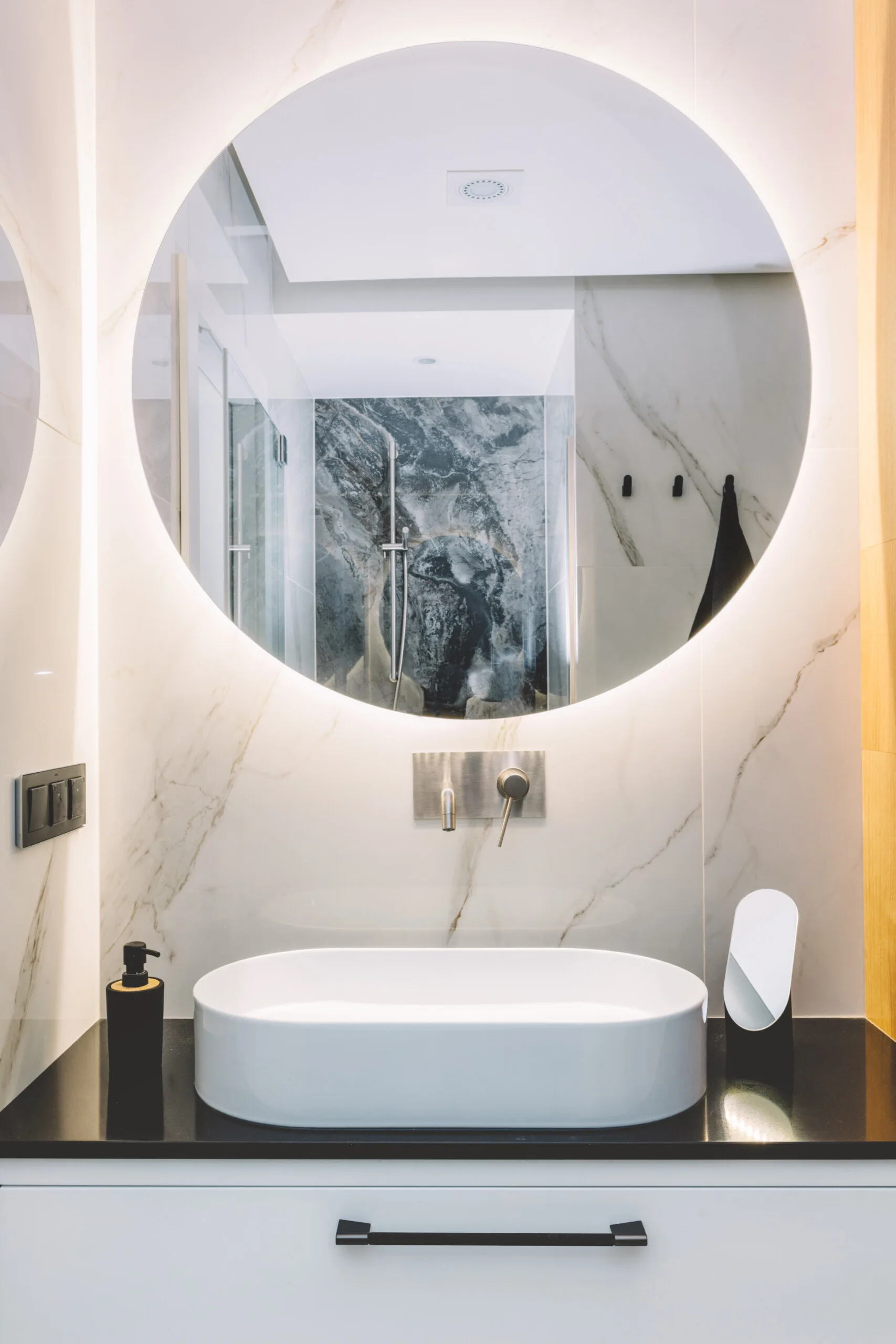 A white ceramic vessel sink sits on a black countertop beneath a large backlit circular mirror mounted on a white marble tile wall.
