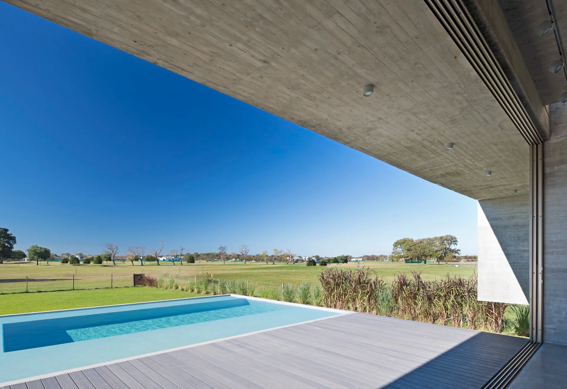 A grey composite deck and rectangular swimming pool sit under a massive raw concrete architectural overhang with an expansive green lawn in the background.
