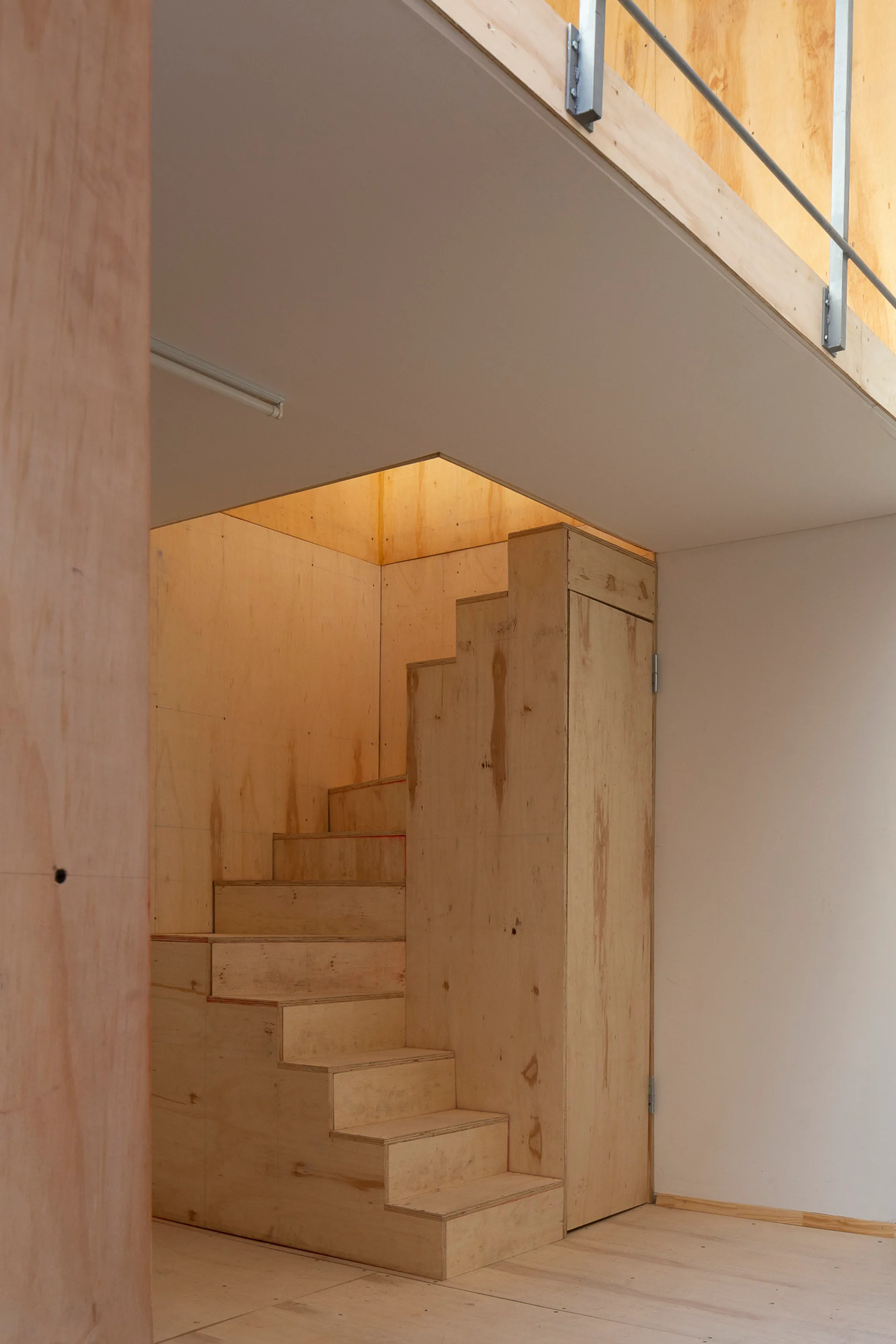 A minimalist staircase built from raw plywood panels leads up to a mezzanine level featuring a thin metal railing against white and wood walls.