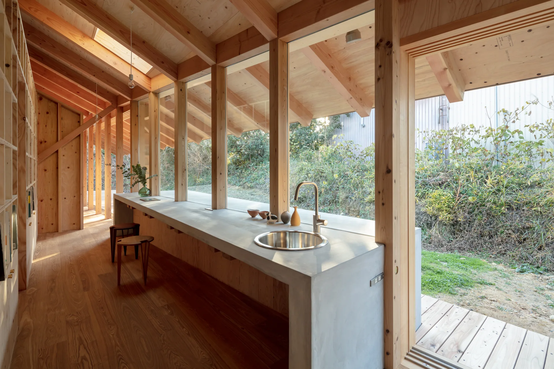 A minimalist kitchen features a long concrete countertop with an integrated circular sink set against a wall of large windows and exposed timber framing.