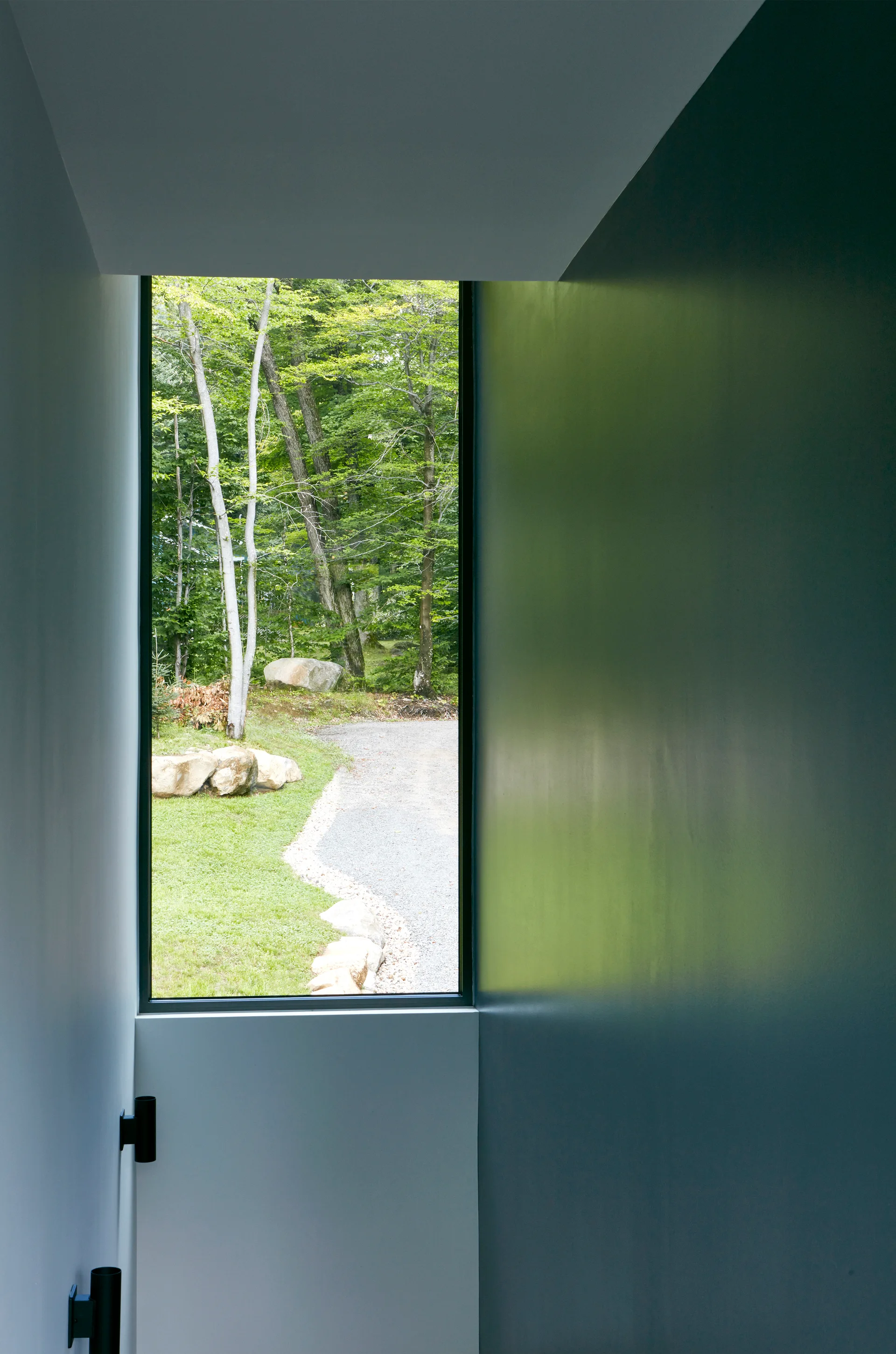 A narrow hallway with dark matte walls leads to a vertical rectangular window showcasing a lush green forest view outside.