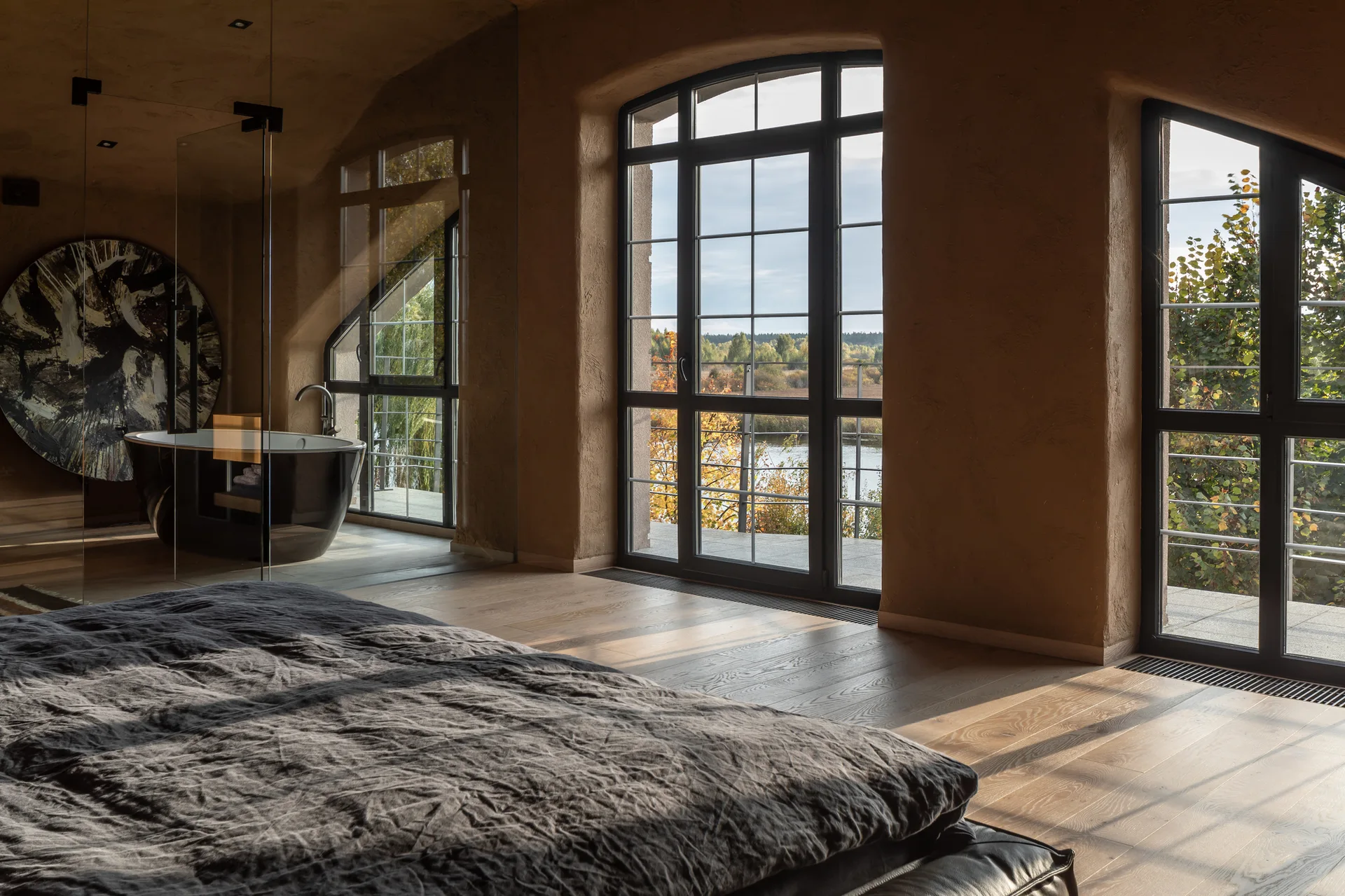 A rustic bedroom features textured earth-toned plaster walls, wood floors, large arched windows, and a black freestanding bathtub behind a glass partition.