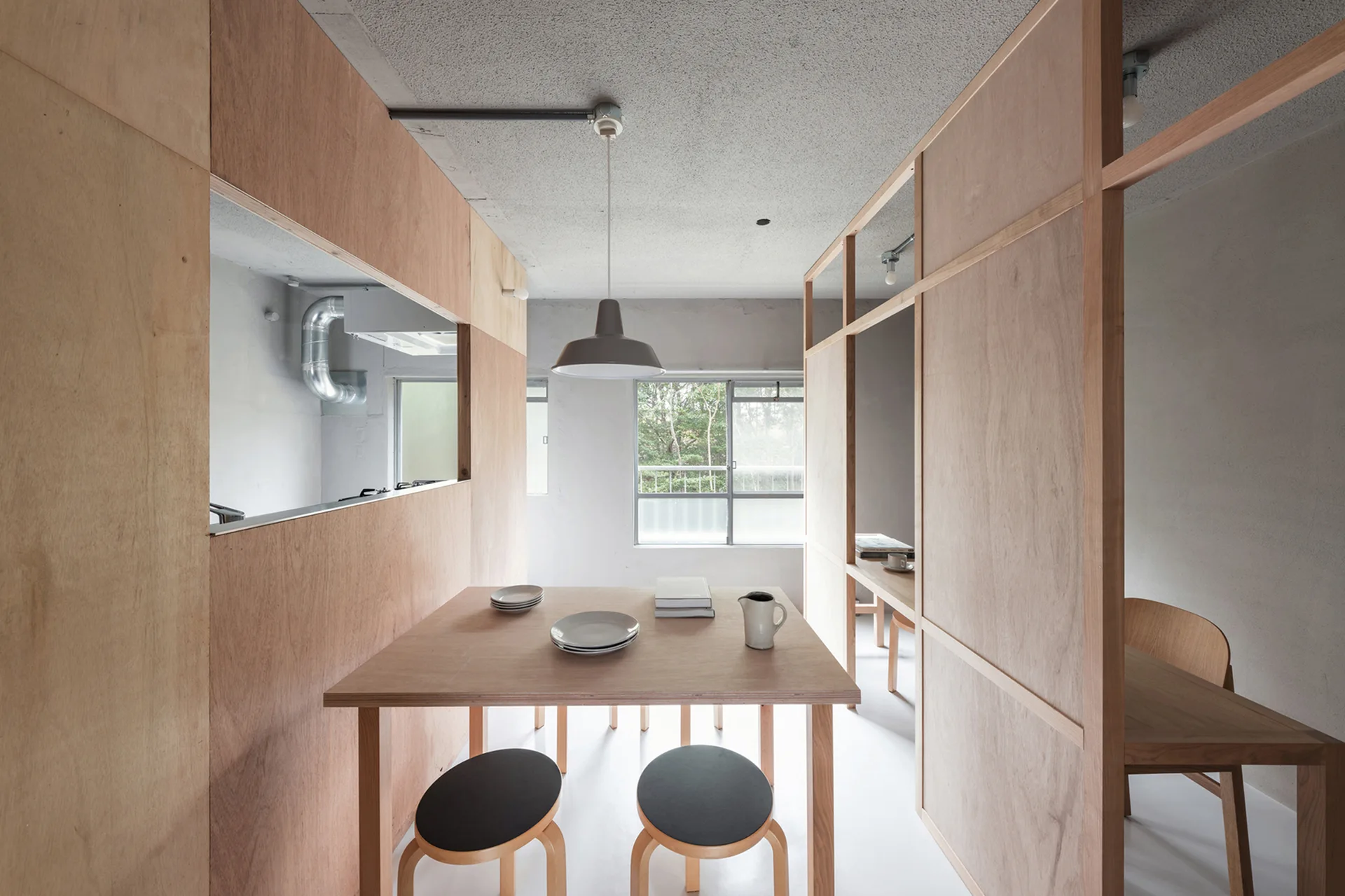A minimalist dining room features a light wood table, stools, a grey pendant light, and plywood wall partitions under a textured concrete ceiling.