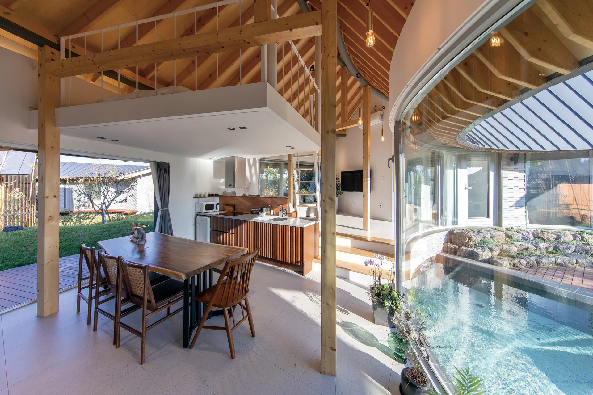 An open-concept dining area with a wood table next to a curved indoor pool, featuring a mezzanine loft overhead and exposed timber rafters.