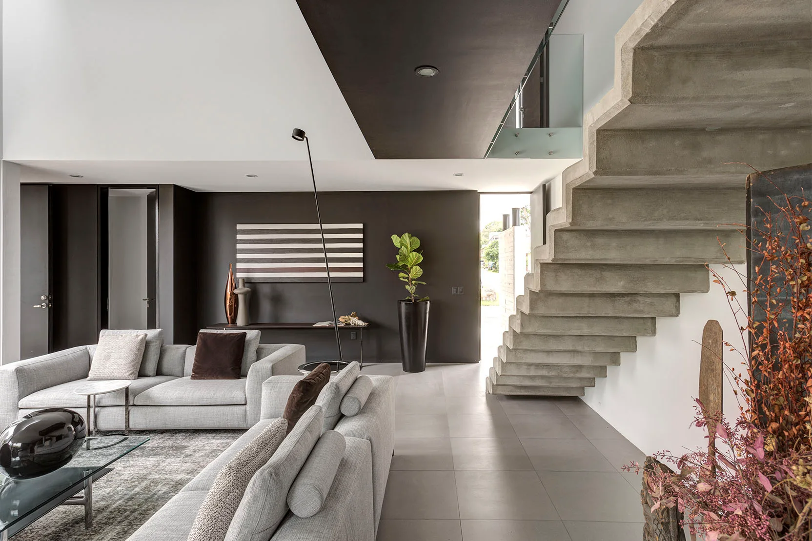 A modern living room features a floating concrete staircase, grey sectional sofas, and a dark charcoal accent wall with minimalist decor.