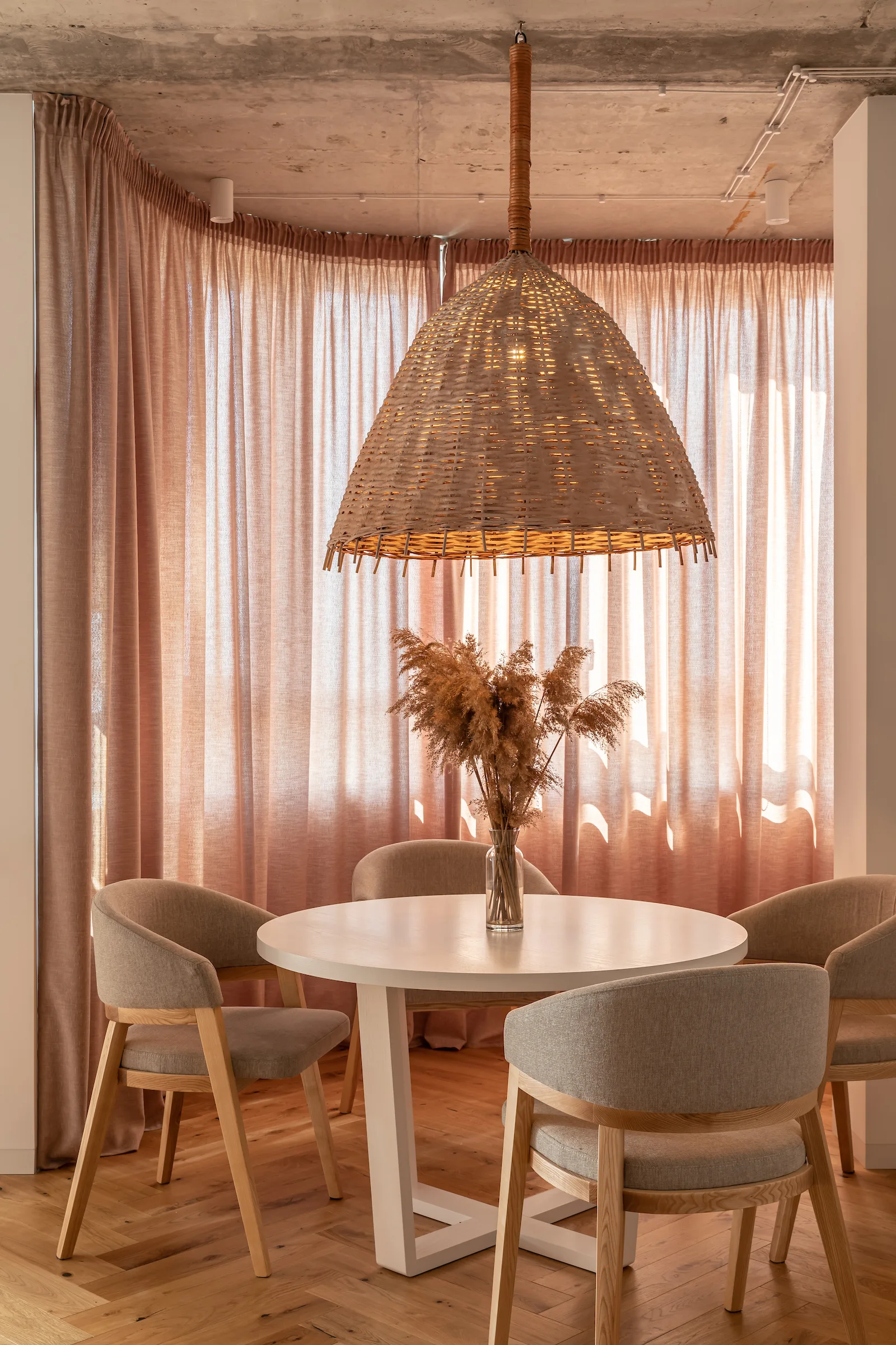 A round white dining table and four grey upholstered chairs sit beneath a large wicker pendant light in front of tall pink curtains.