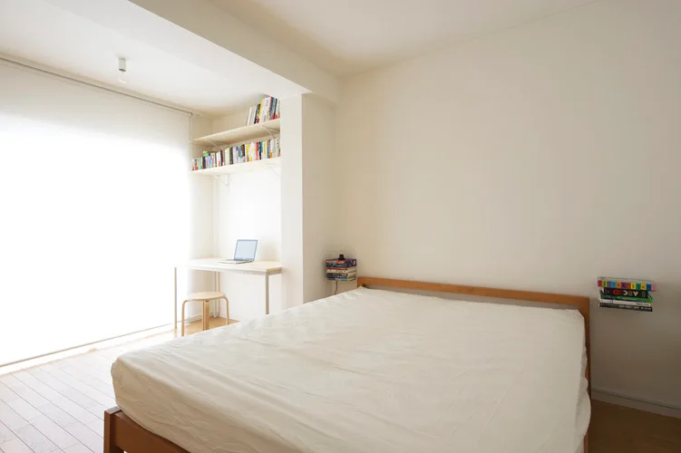 A minimalist white bedroom features a light wood bed, a desk tucked into a corner alcove, and floating shelves filled with books.