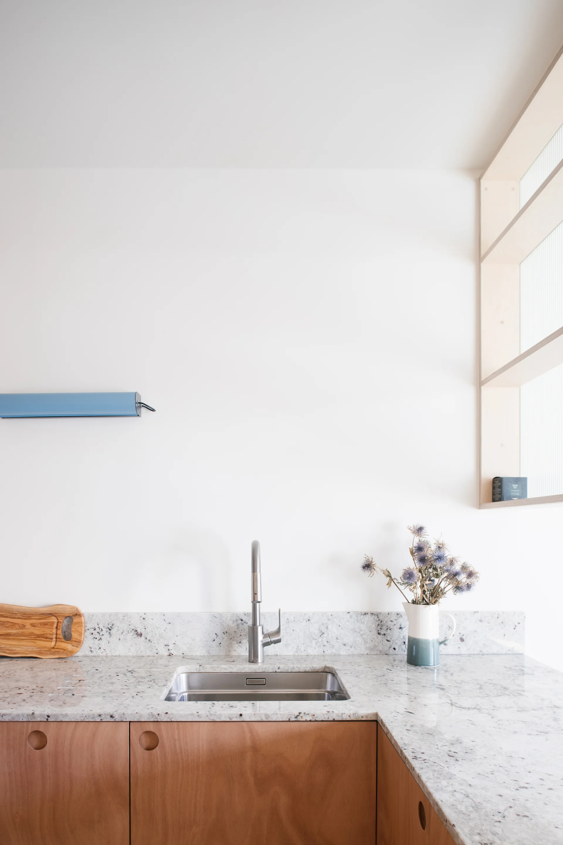 A minimalist kitchen corner featuring wood base cabinets, a speckled stone countertop with a stainless steel sink, and a clean white wall.
