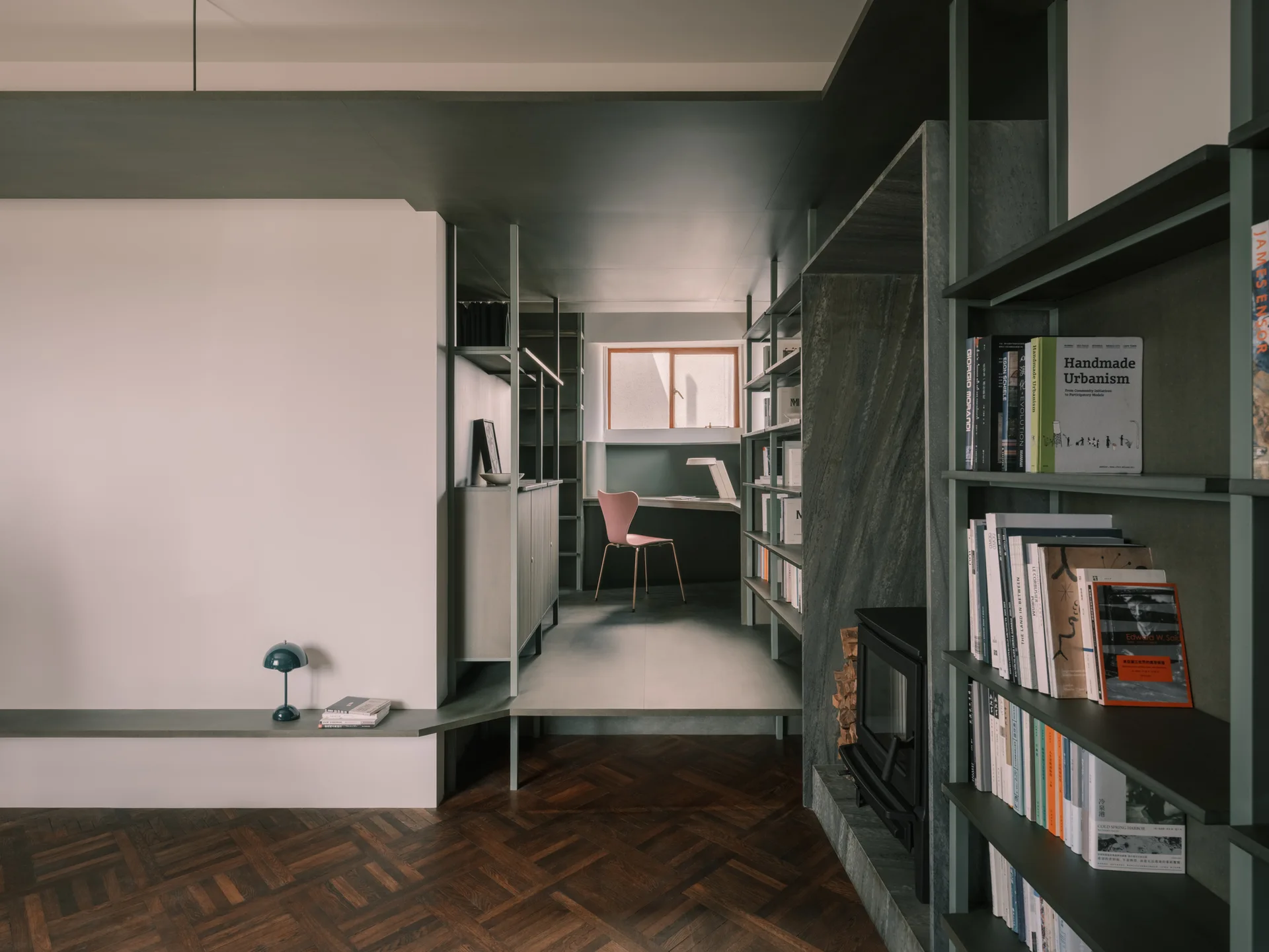 A modern home office features sage green built-in shelving, a raised platform desk area, a pink chair, and dark herringbone wood floors.