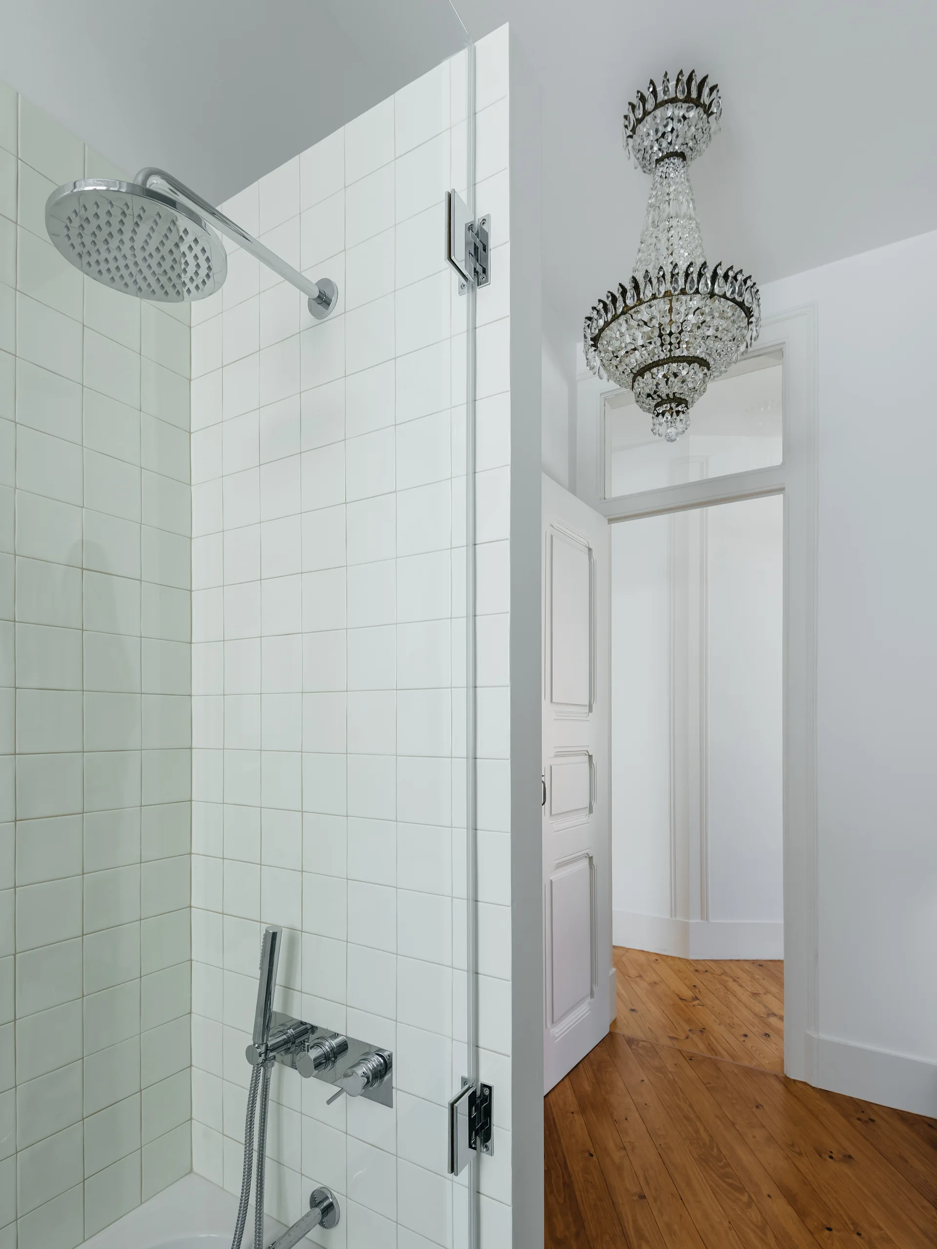 A white tiled shower enclosure with chrome hardware stands next to an open doorway showing wood floors and a hanging crystal chandelier.