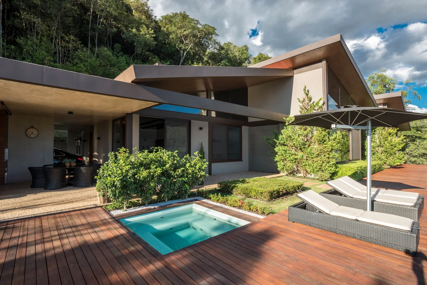 A modern residential backyard featuring a wide wood plank deck, a small square built-in pool, and two black wicker lounge chairs with a dark umbrella.