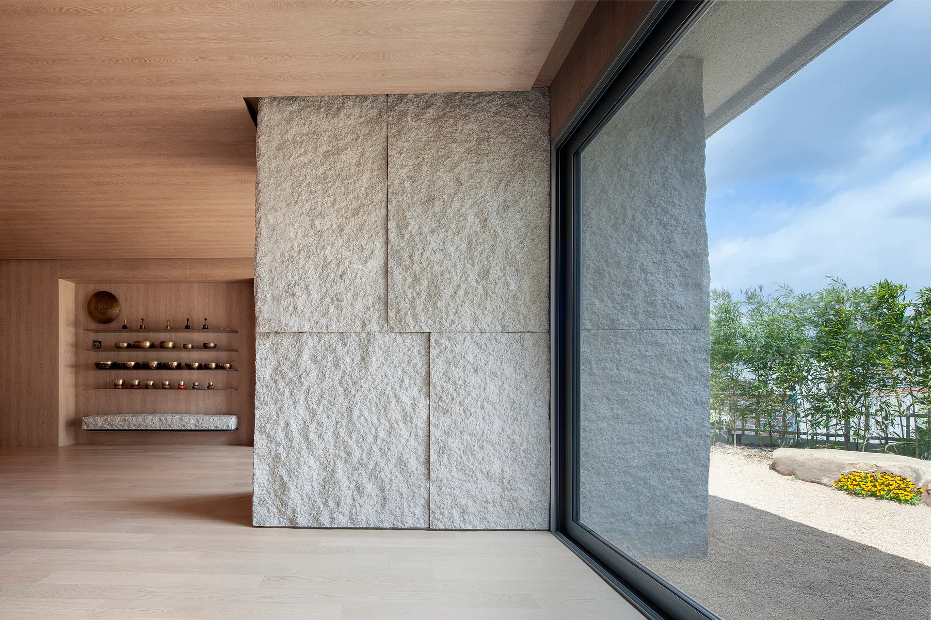A minimalist meditation room featuring light wood paneling, a textured stone accent wall, and a large sliding glass door looking out to a garden.