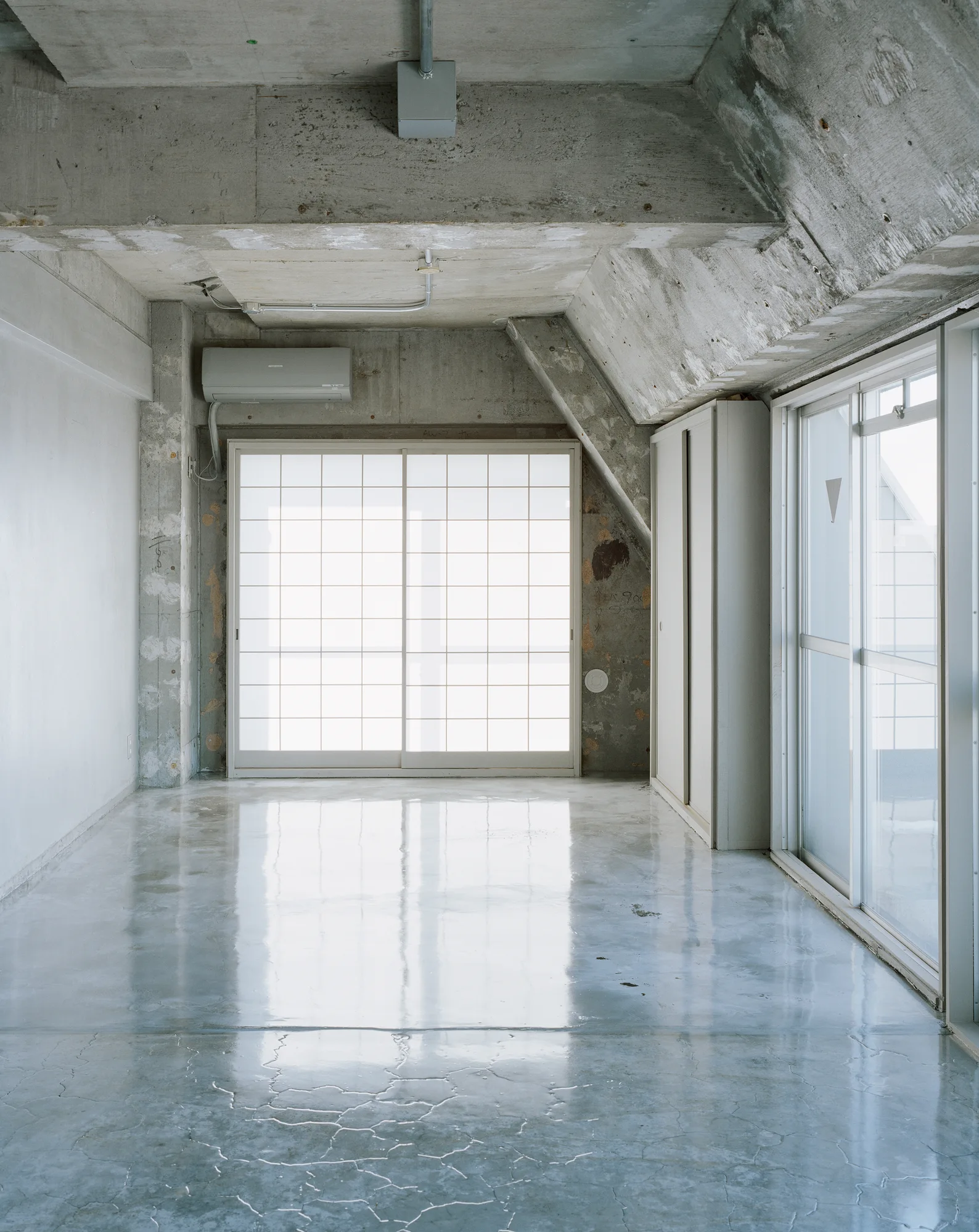 An empty interior room features a highly reflective polished concrete floor, raw concrete ceiling, and a translucent grid shoji-style sliding partition.
