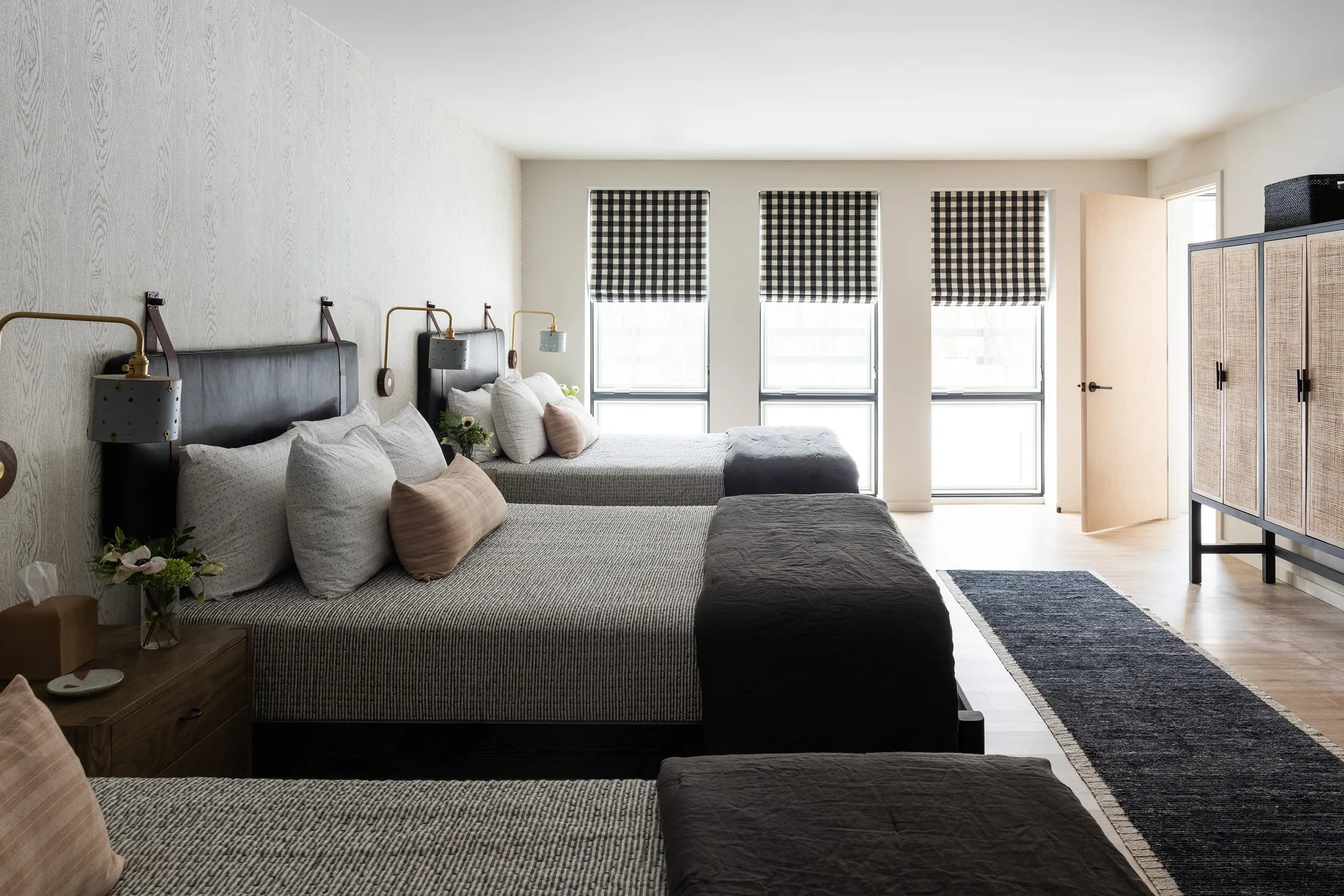 A shared bedroom features three beds in a row against textured wallpaper, with three narrow windows dressed in black-and-white checkered roman shades.