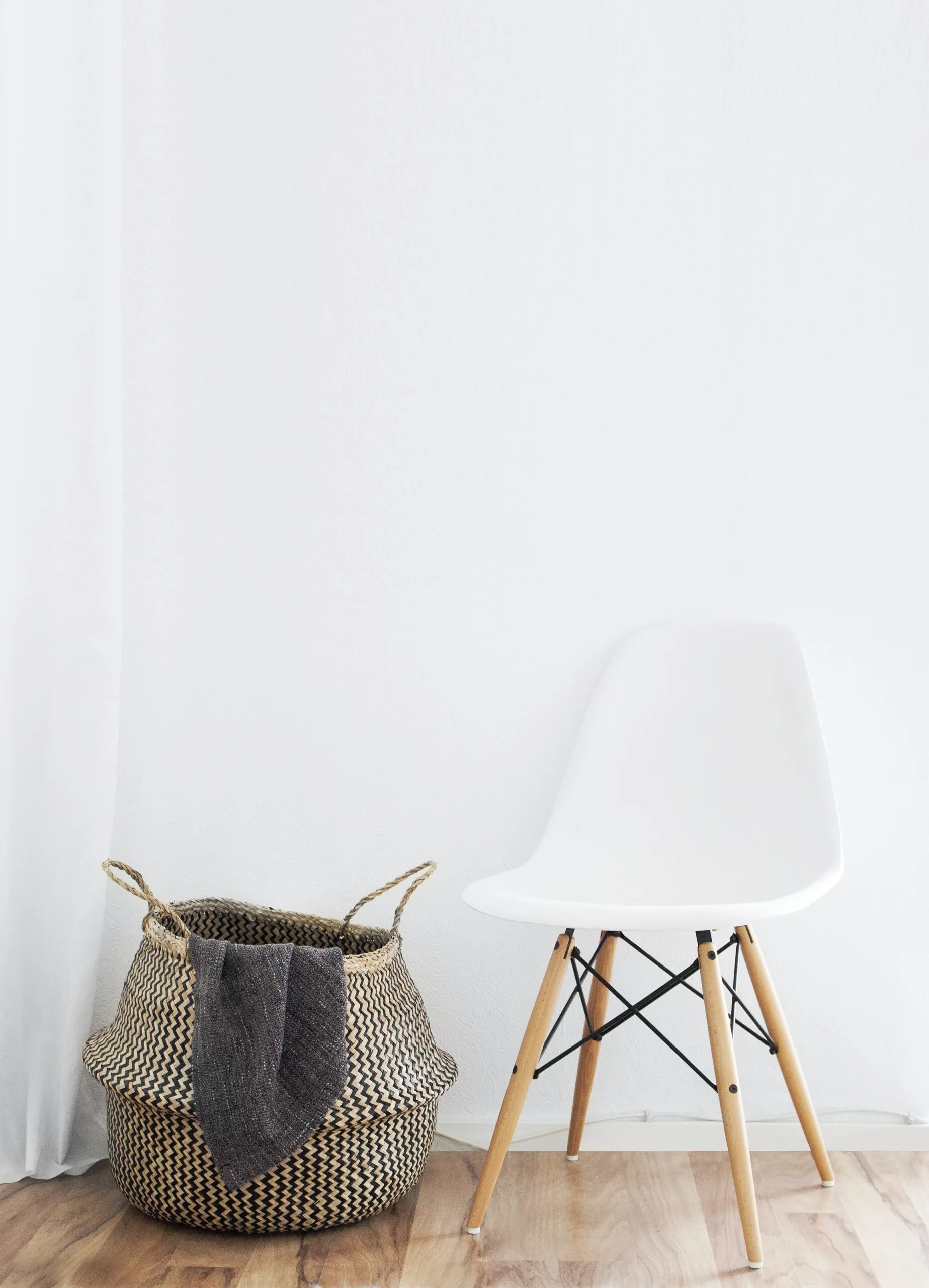 A white molded plastic chair with wooden legs sits next to a black-and-white chevron woven basket containing a grey throw on a light hardwood floor.