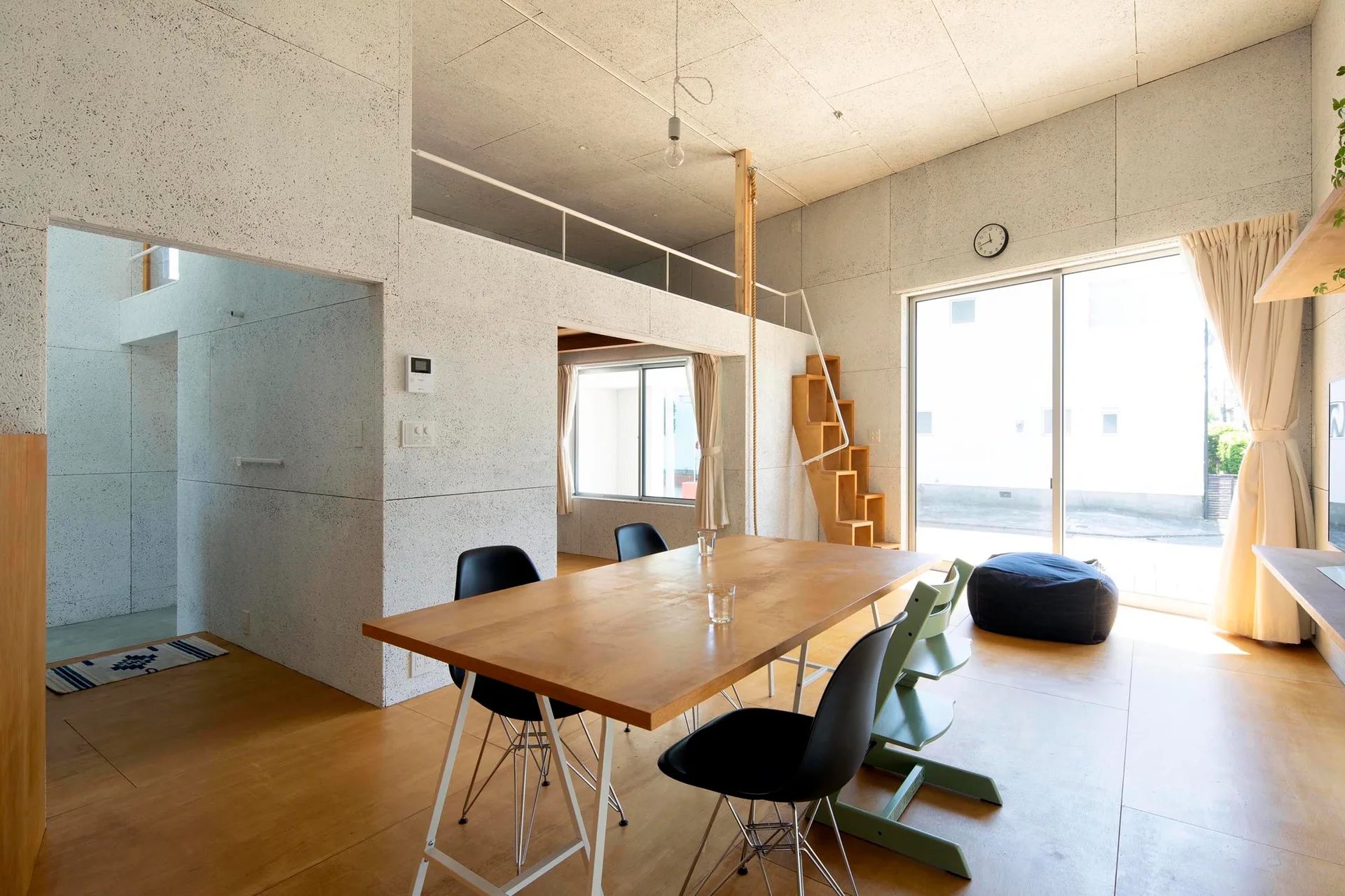 A minimalist dining space featuring speckled composite walls, a wood dining table with chairs, and a wooden alternating-tread staircase leading to a loft.