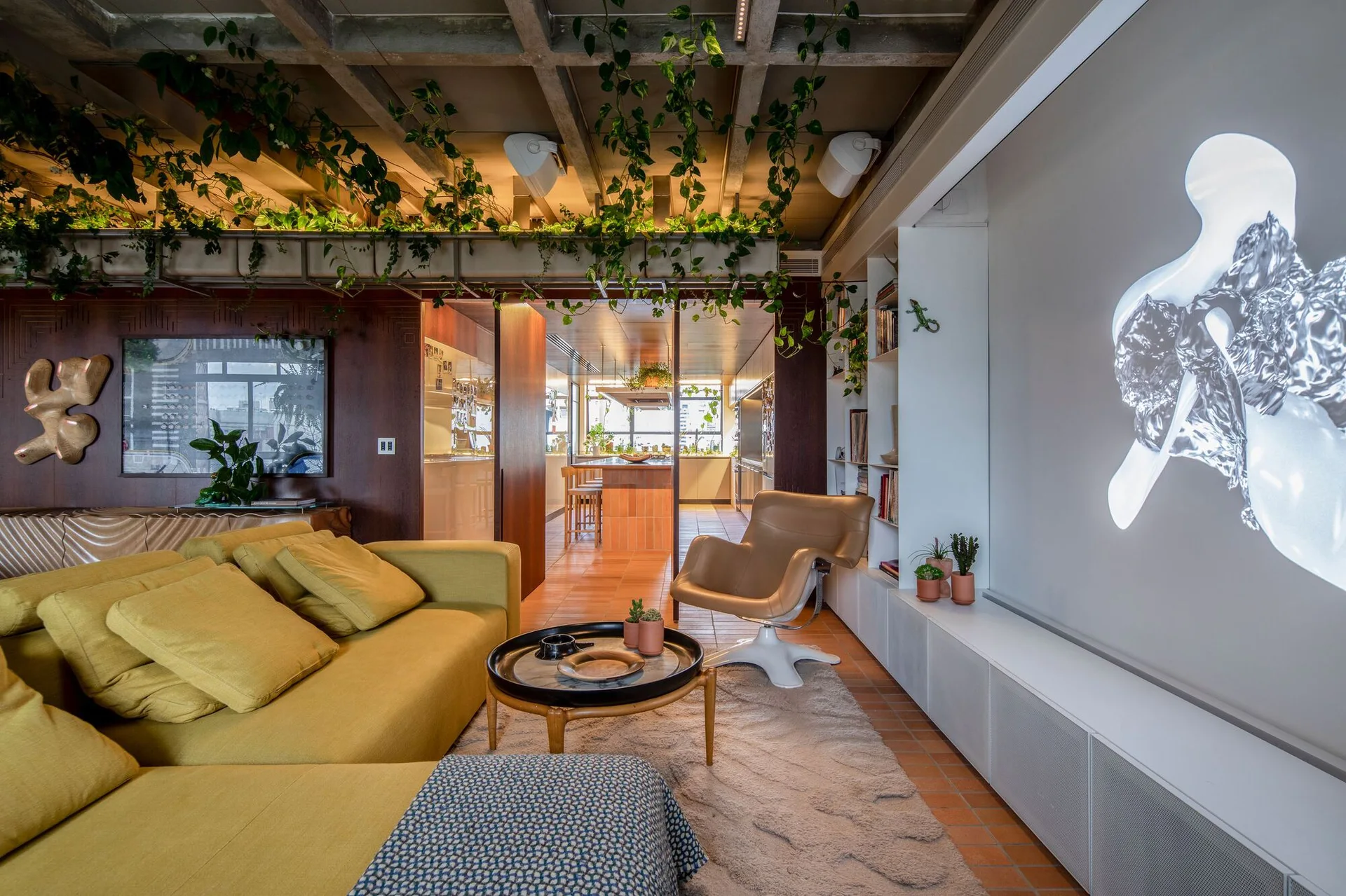 A living room features a yellow sectional sofa, a mid-century swivel armchair, and an exposed concrete grid ceiling draped with hanging green plants.