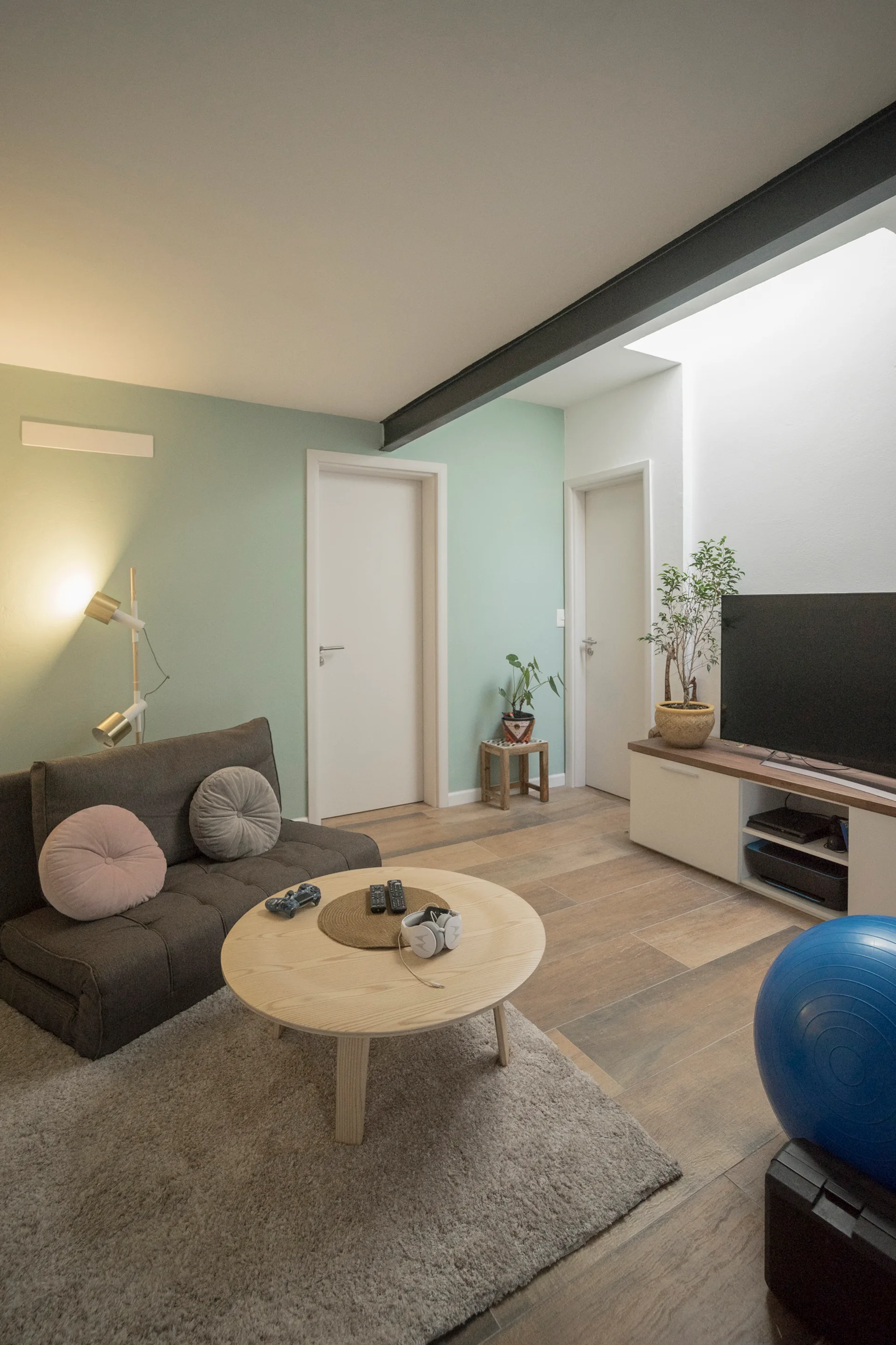A modern living room features a dark grey sofa, round wood coffee table, and a media console set against mint green walls under a dark ceiling beam.