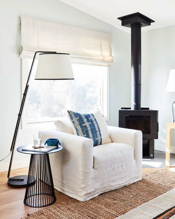 A white slipcovered armchair with a blue patterned pillow sits next to a black metal wood stove and a slender floor lamp.