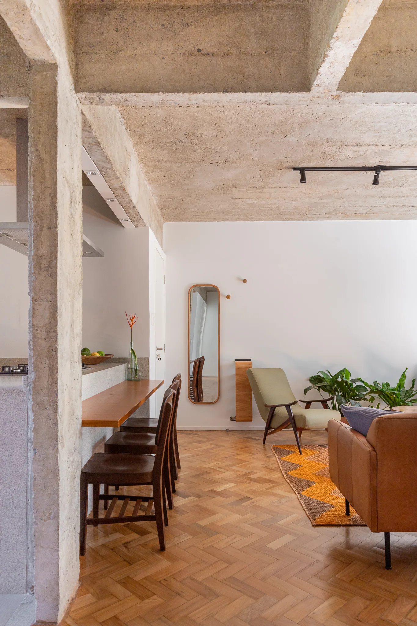 An open plan living area features an exposed concrete ceiling, herringbone wood parquet flooring, a green armchair, and a wood bar counter with stools.