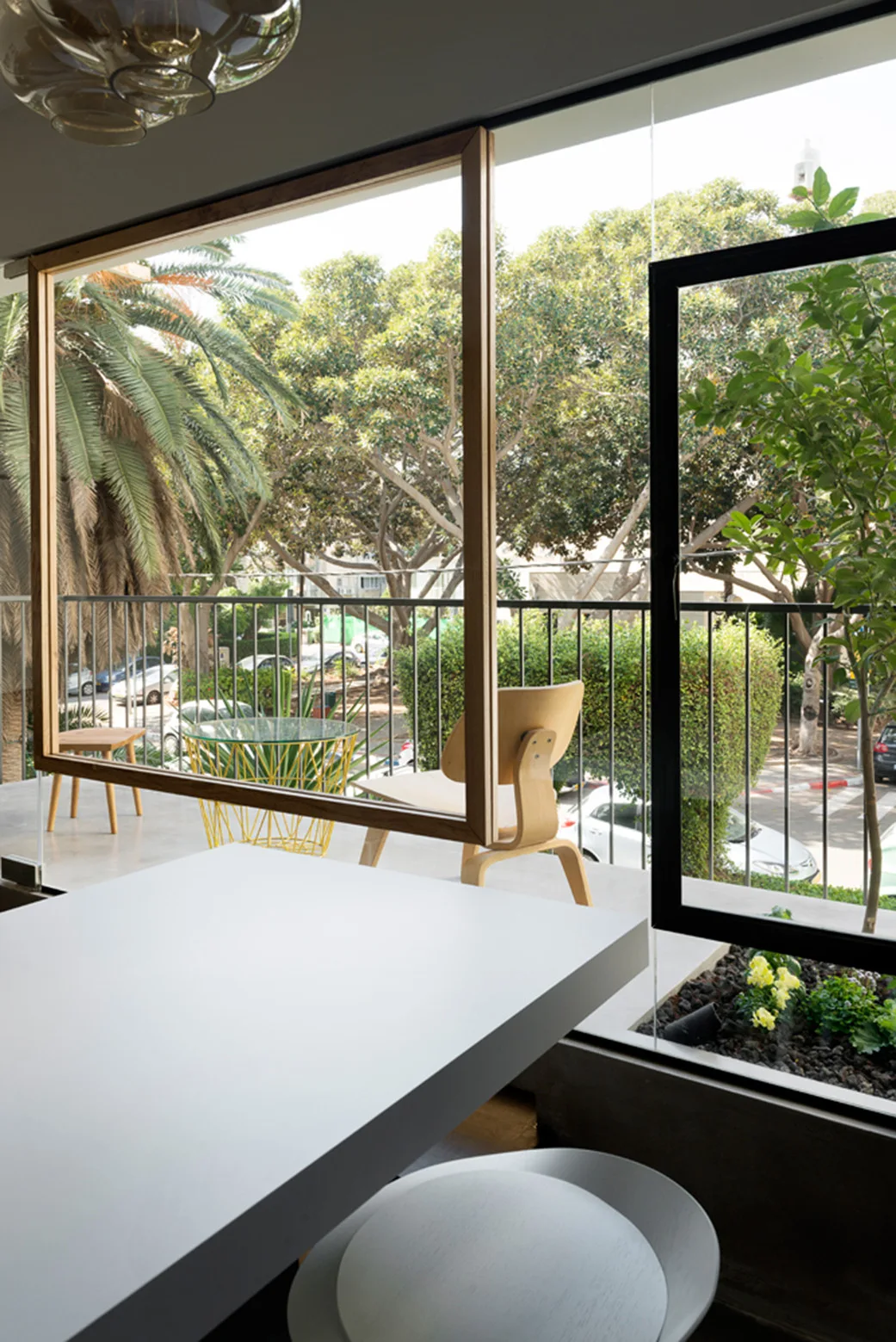 A white dining table looks out through a large glass window to a balcony with a wooden chair and a yellow wire side table.
