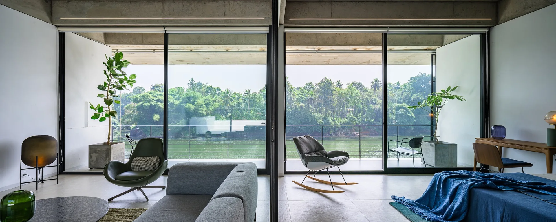 A modern open-concept space featuring an exposed concrete ceiling, minimalist furniture, and large sliding glass doors opening to a river view balcony.