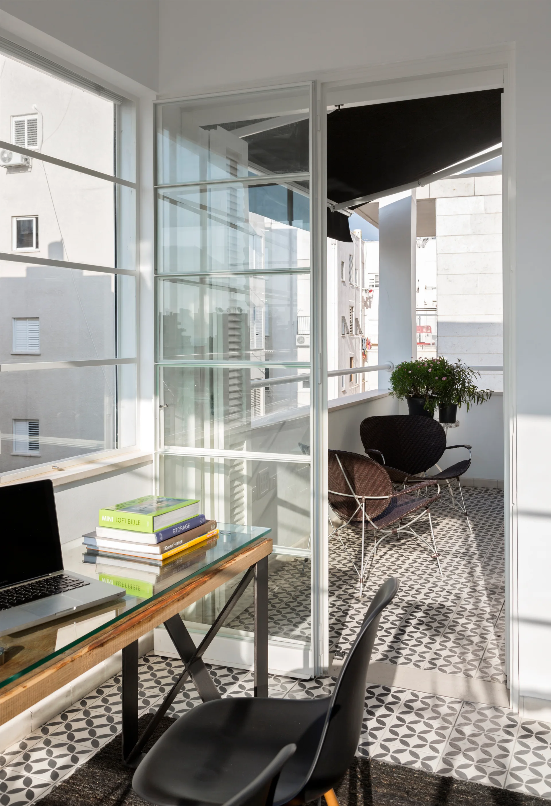 A modern home office with a glass desk opens onto a balcony with patterned tiles, outdoor chairs, and a black awning.
