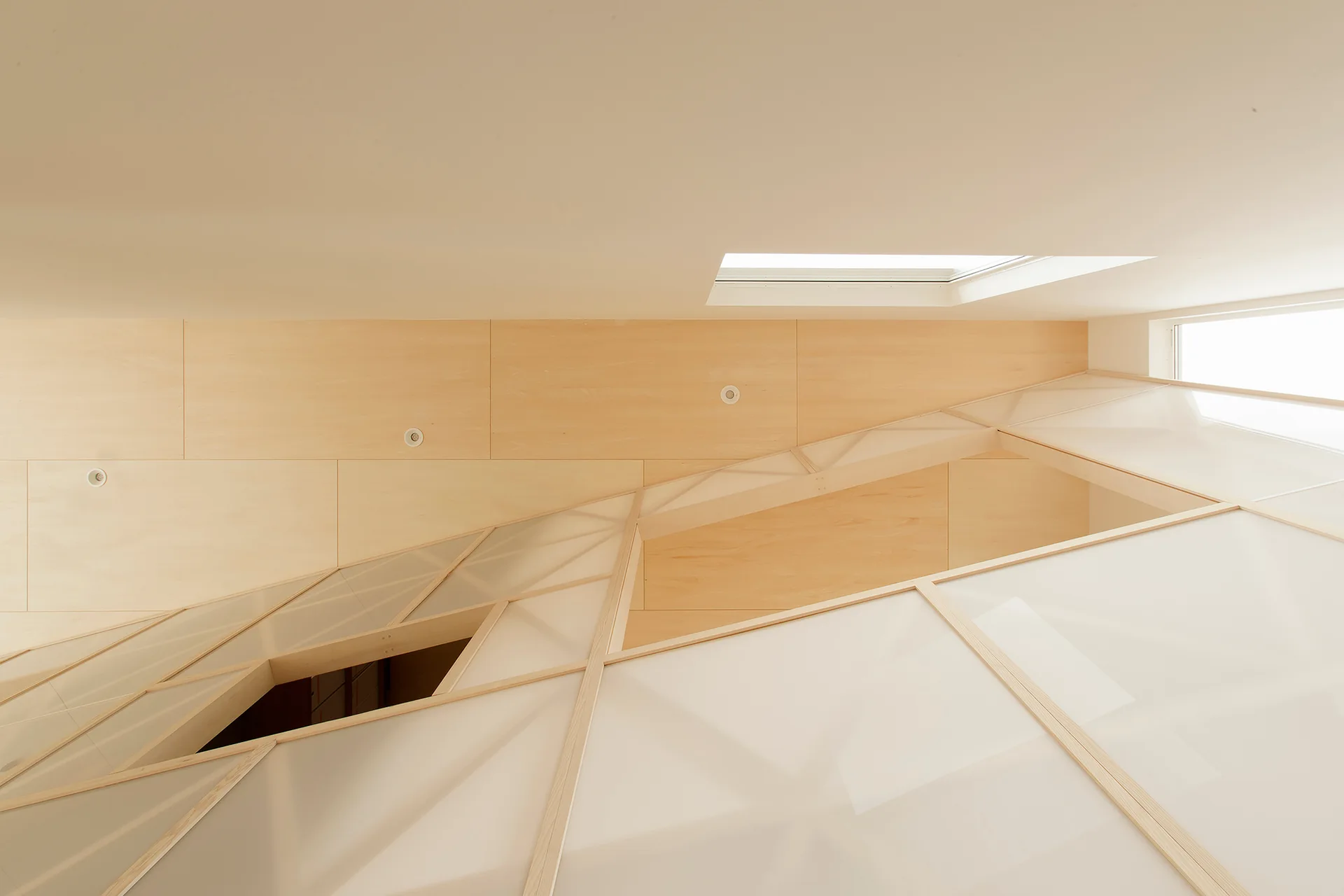 A modern stairwell ceiling features light wood panels, a small skylight, and a geometric screen made of frosted panels in light timber framing.