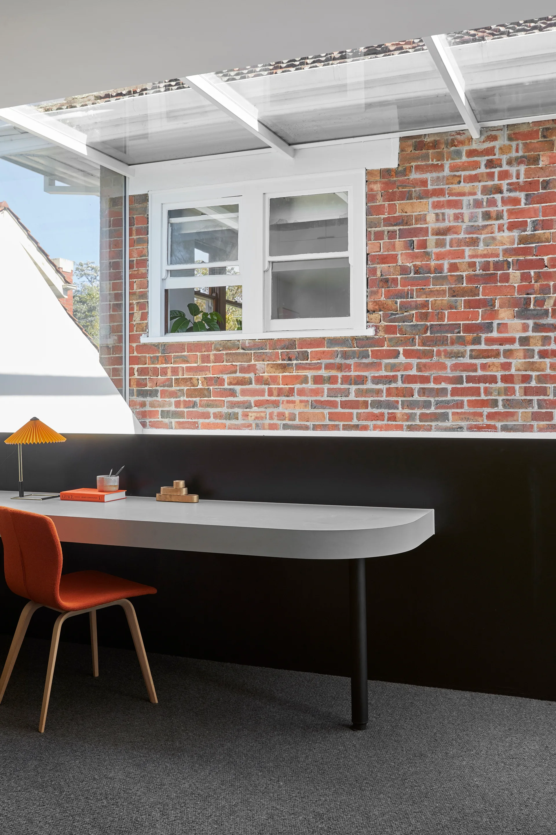 A modern gray desk with an orange chair is positioned against a black wall panel beneath an exposed red brick wall and glass skylight.