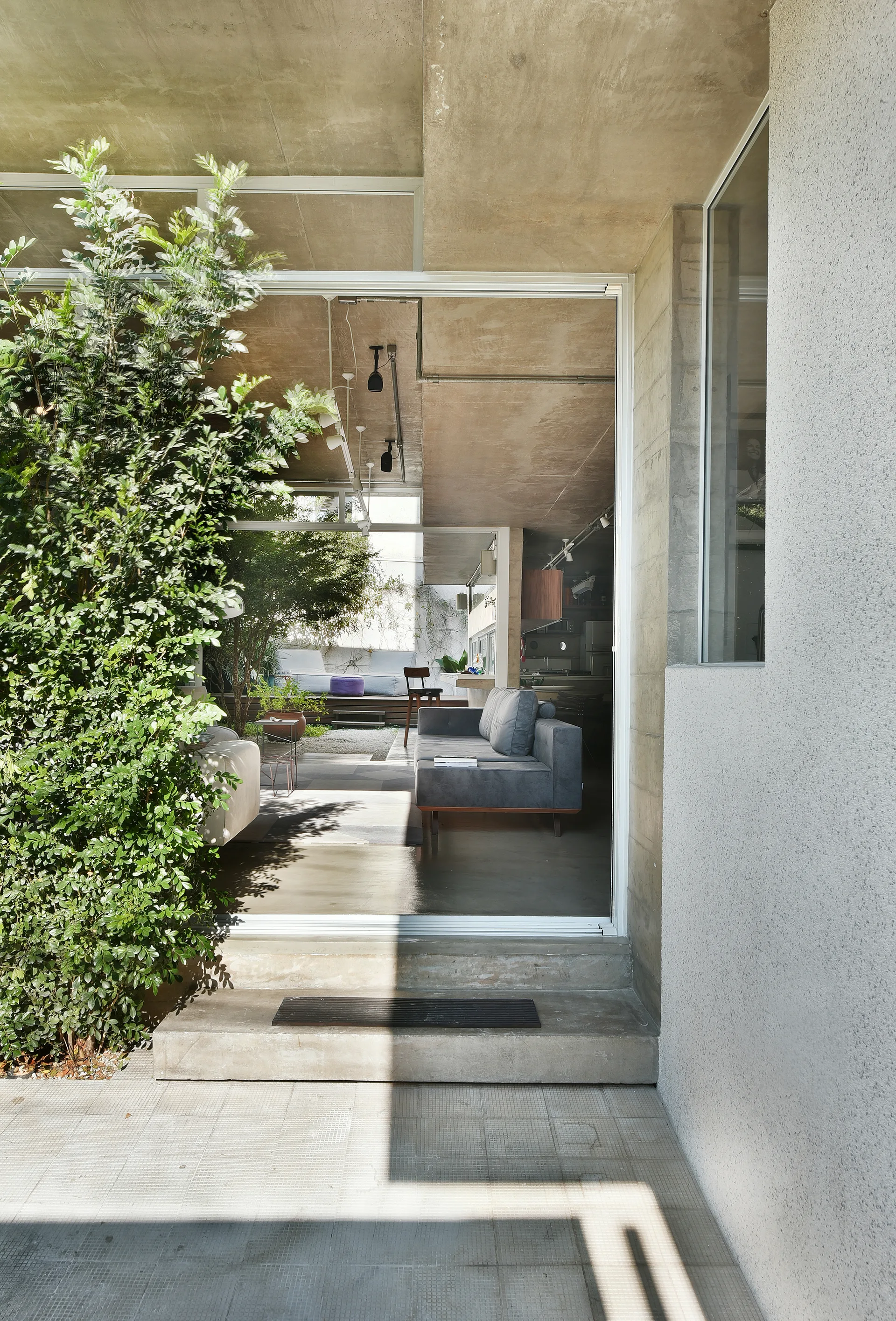 Looking through a large glass sliding door from a concrete patio into a modern living room with a grey sofa and exposed concrete ceiling.