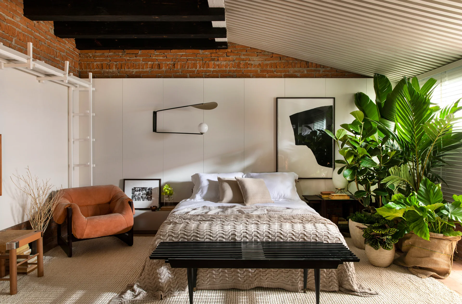 An industrial loft bedroom features a bed with a knit coverlet, a cluster of green potted plants, and an upper wall of exposed red brick.