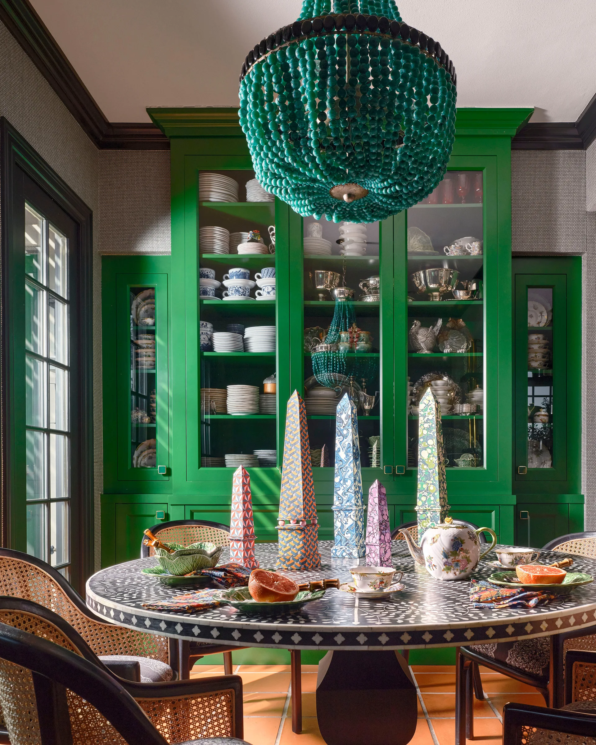 A round patterned bone inlay dining table surrounded by cane-back chairs sits in front of a tall, vibrant green display cabinet filled with china.