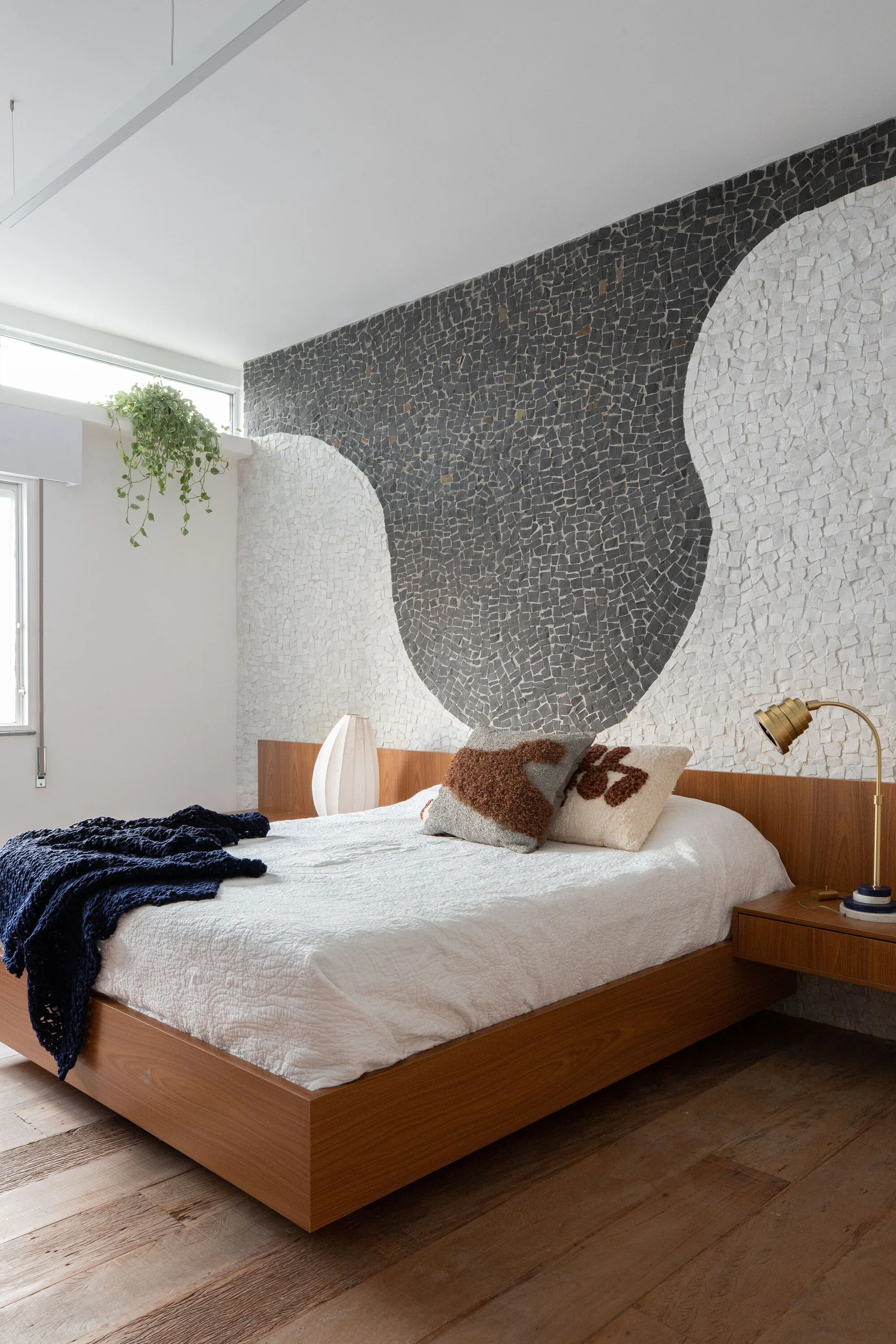 A modern bedroom features a wooden platform bed against a black and white wavy stone mosaic accent wall with integrated floating nightstands.