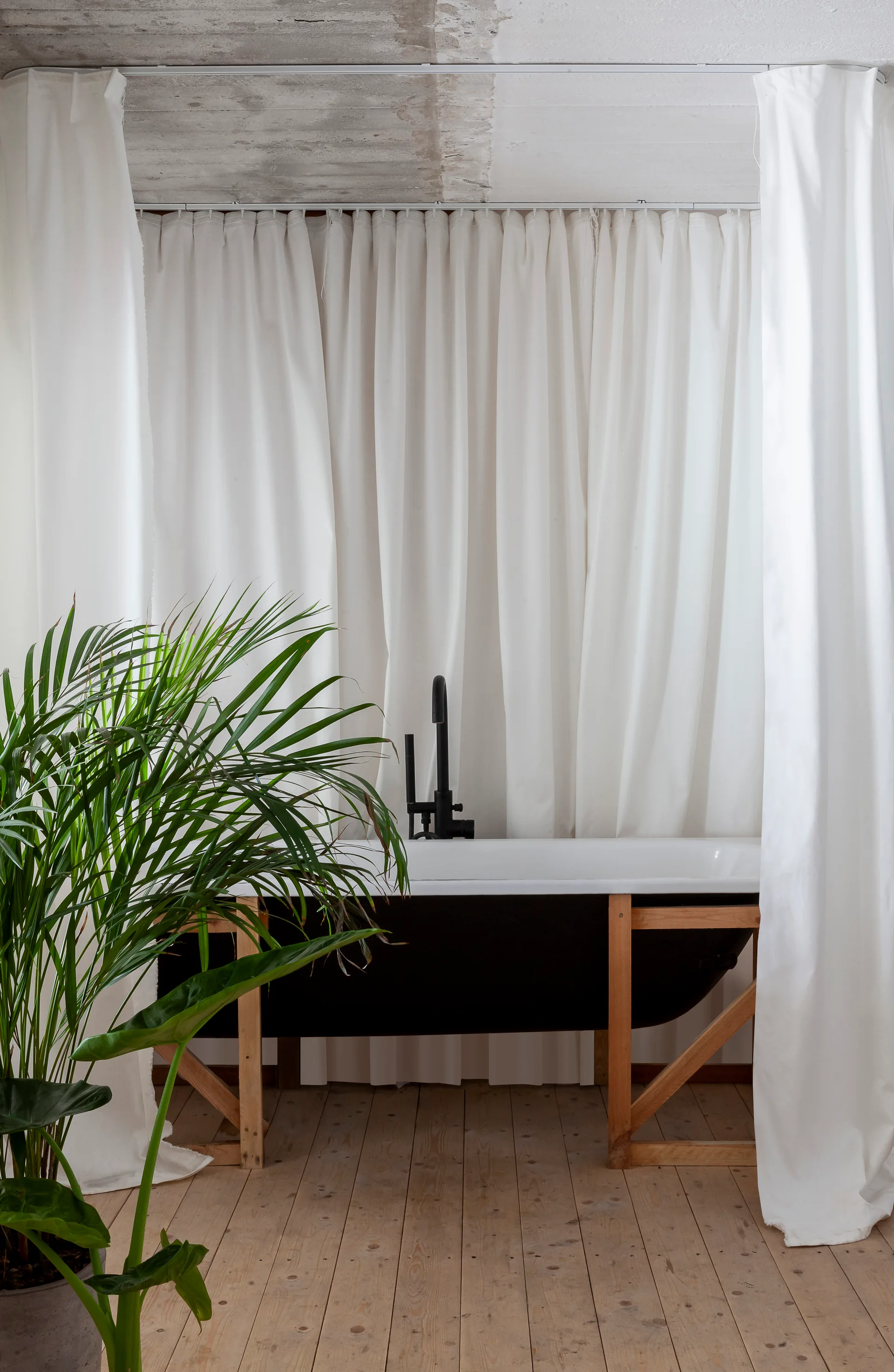 A dark freestanding bathtub supported by a light wood frame sits against a backdrop of full-length white curtains on wood plank floors.