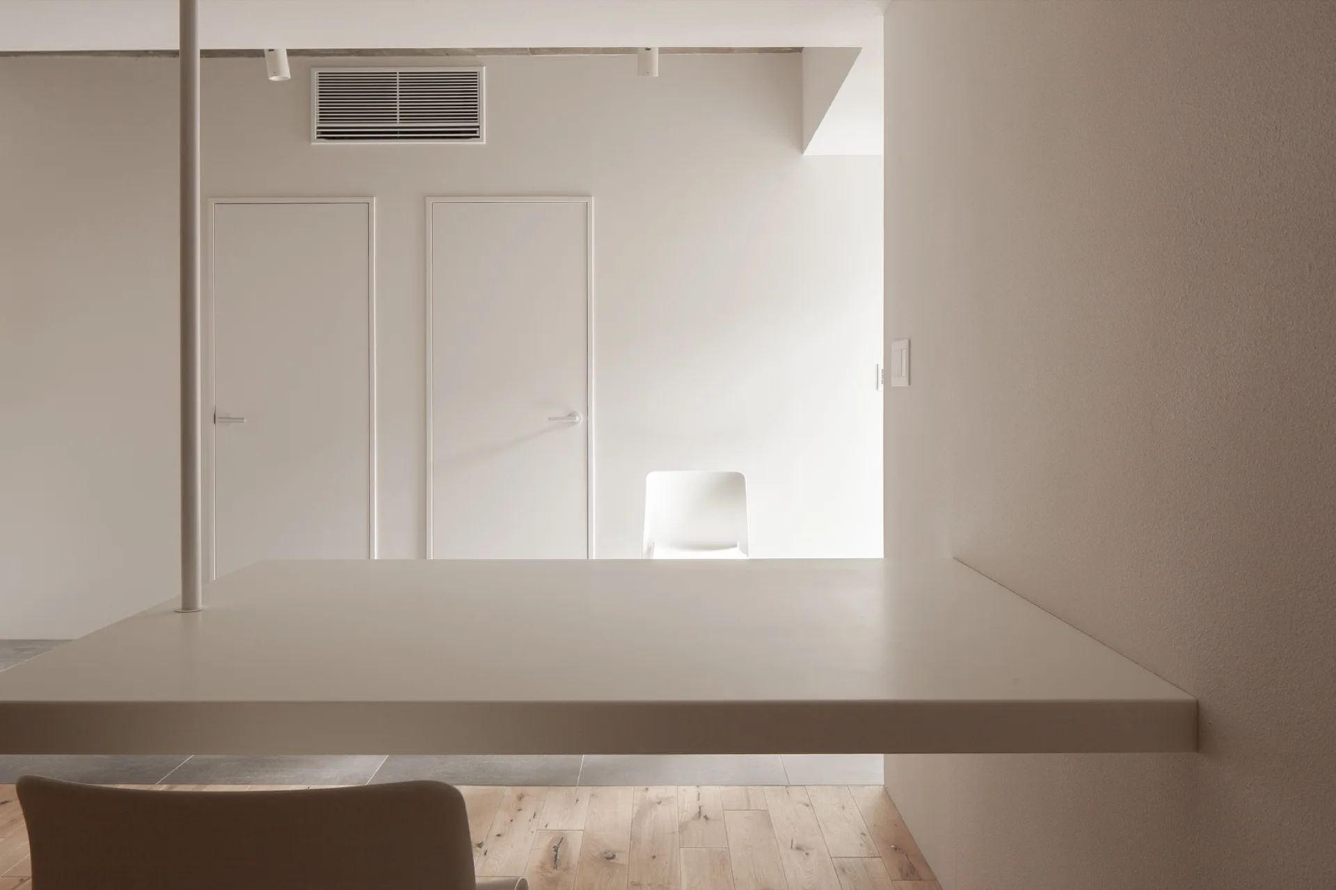 A minimalist white room features a cantilevered table supported by a single thin metal pole, set against light wood flooring and two closed doors.