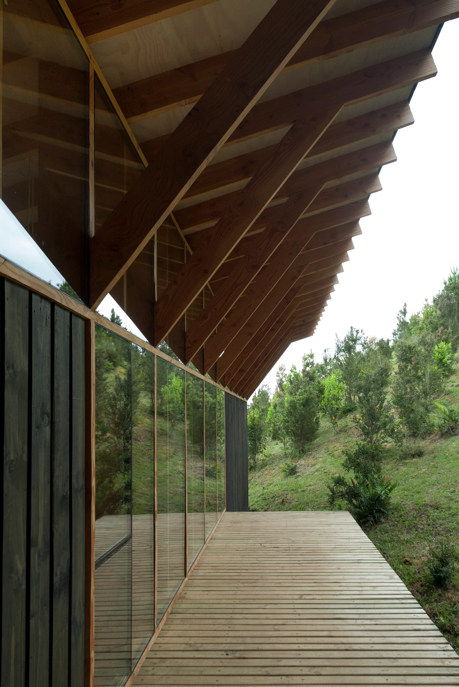 A modern wooden deck with slatted flooring beneath angled timber rafters next to a glass wall reflecting a grassy hillside.