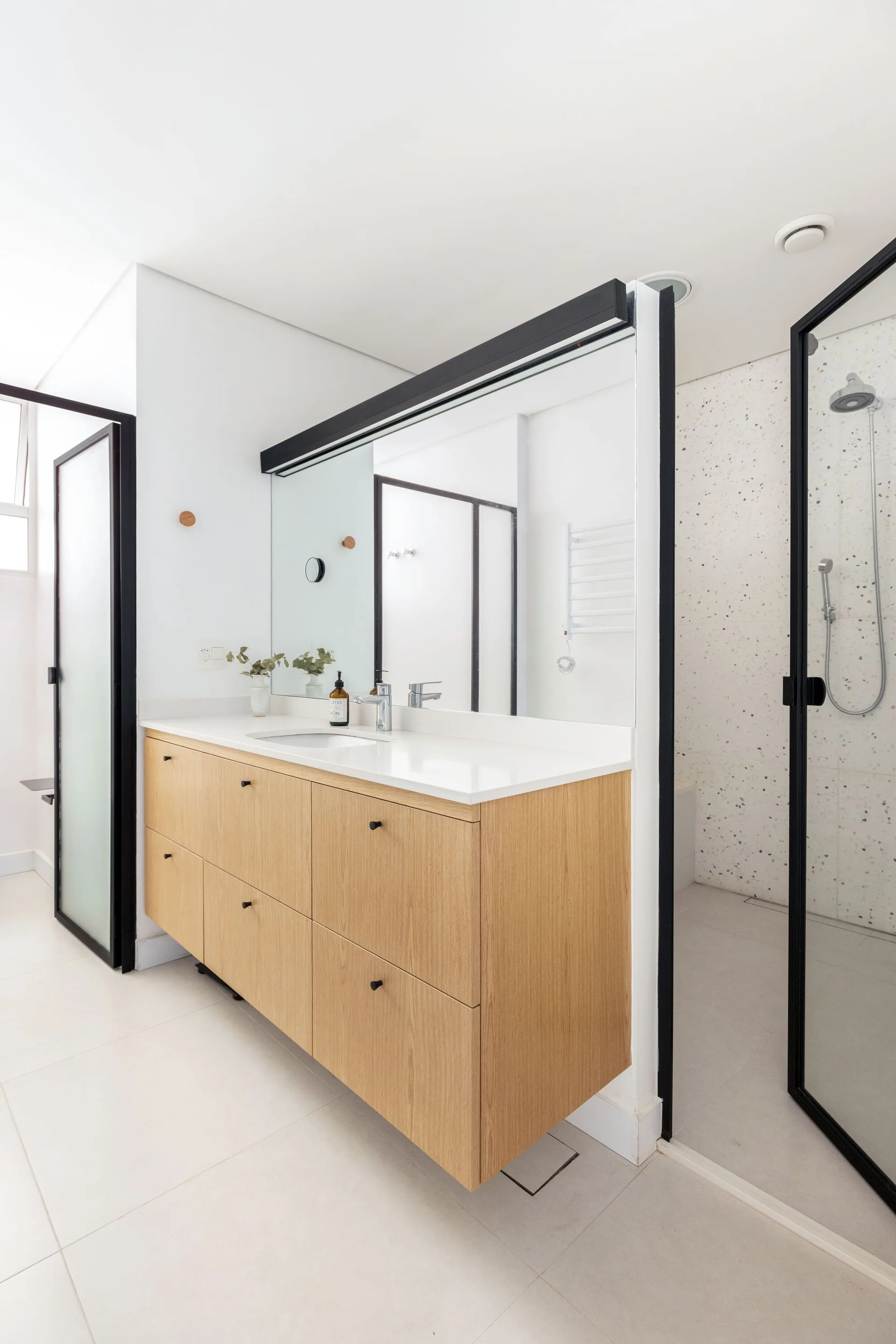 A modern bathroom features a floating wood vanity with a white countertop next to a black-framed glass shower enclosure with speckled tiles.