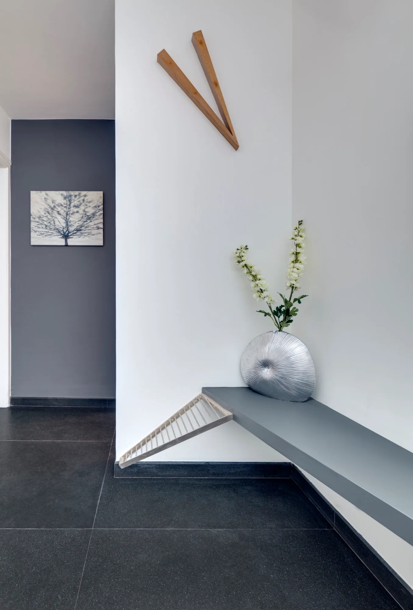 A minimalist entryway corner features a grey floating shelf holding a silver textured vase with white flowers, beneath wooden clock hands on a white wall.