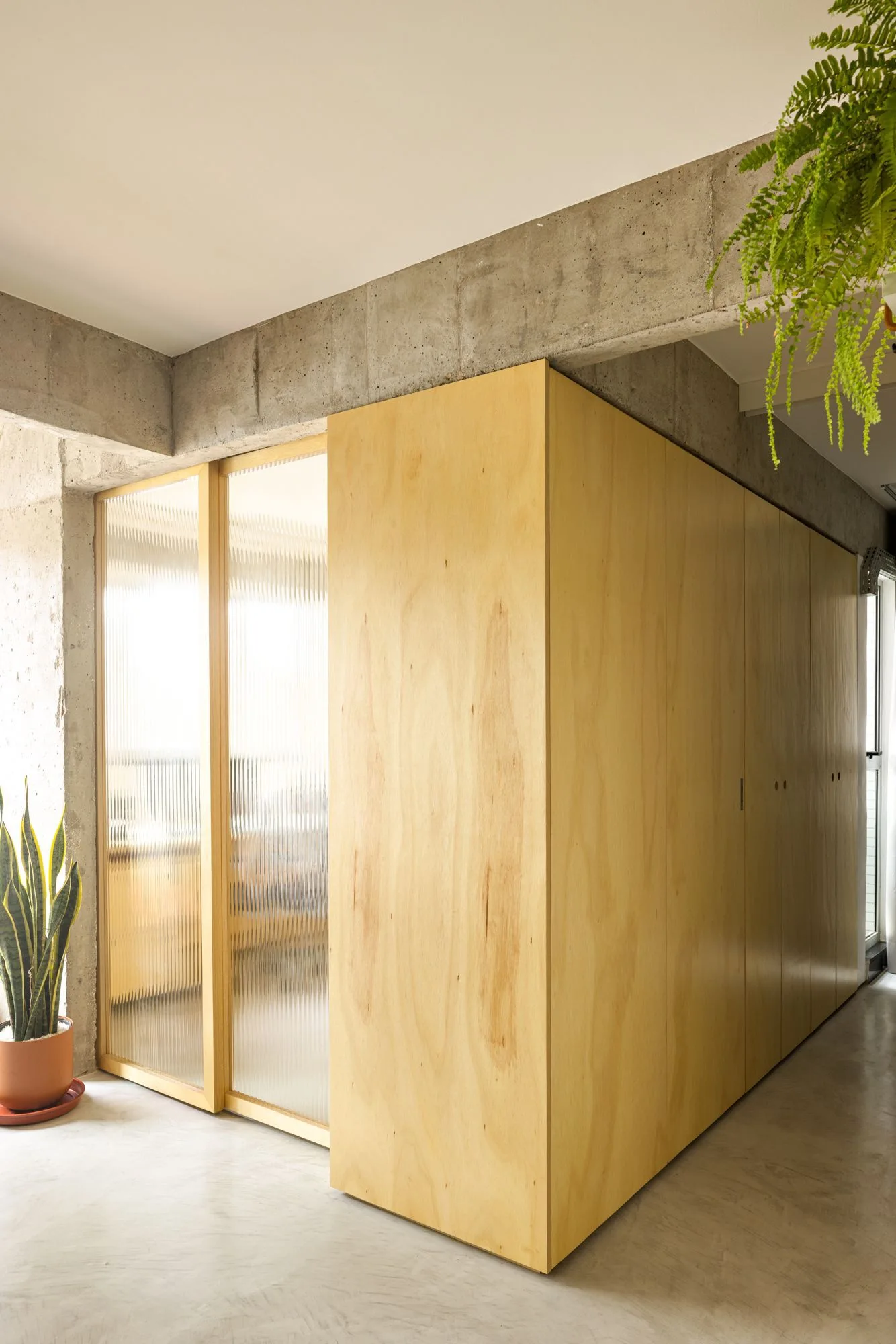 A light plywood cabinet wall stands adjacent to a fluted glass partition beneath an exposed concrete ceiling beam.