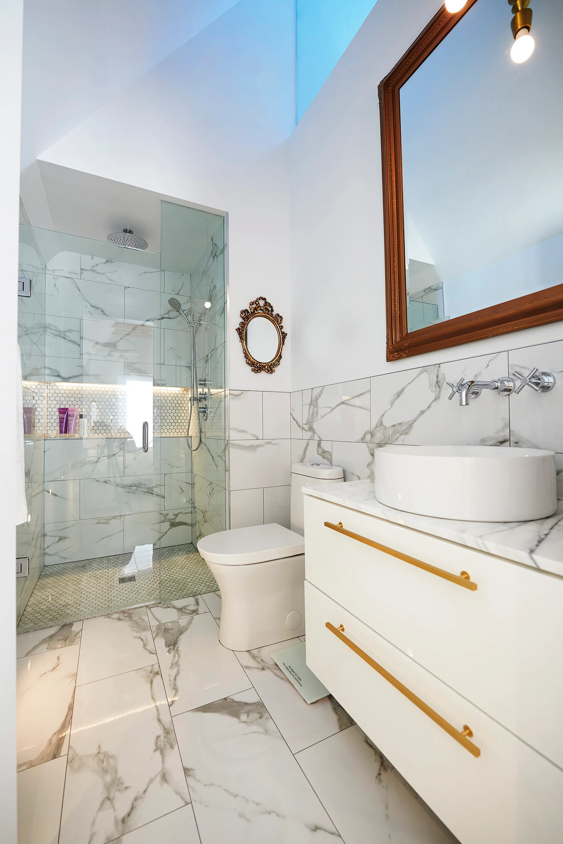 A modern bathroom with white marble-patterned floor and wall tiles, a white vanity with gold handles, a walk-in shower, and a skylight above.